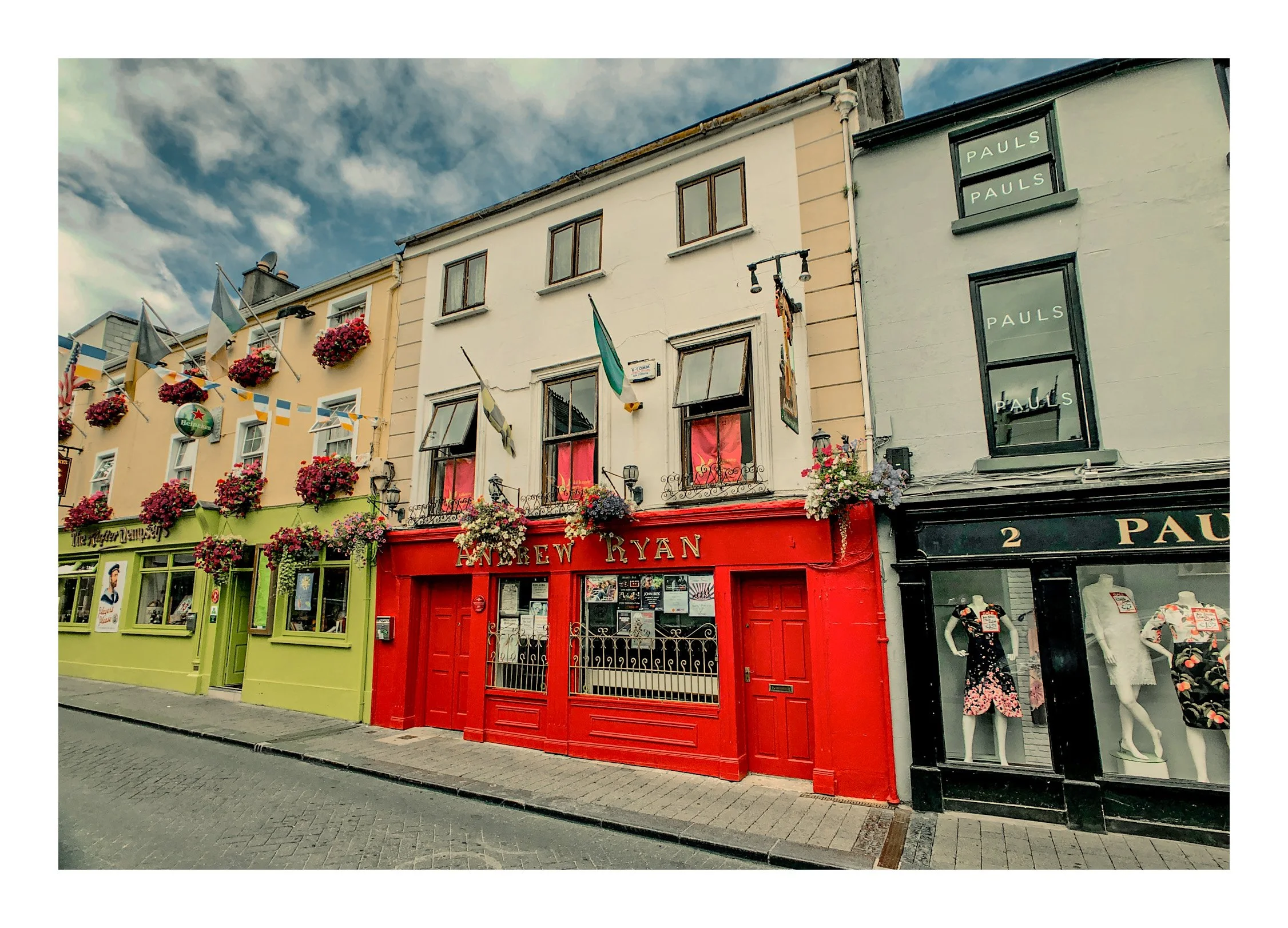 Colorful street scene with three narrow storefronts decorated with flowers and flags. The stores are painted in bright colors: green, red, and black, with posters in the windows. The sky is partly cloudy.