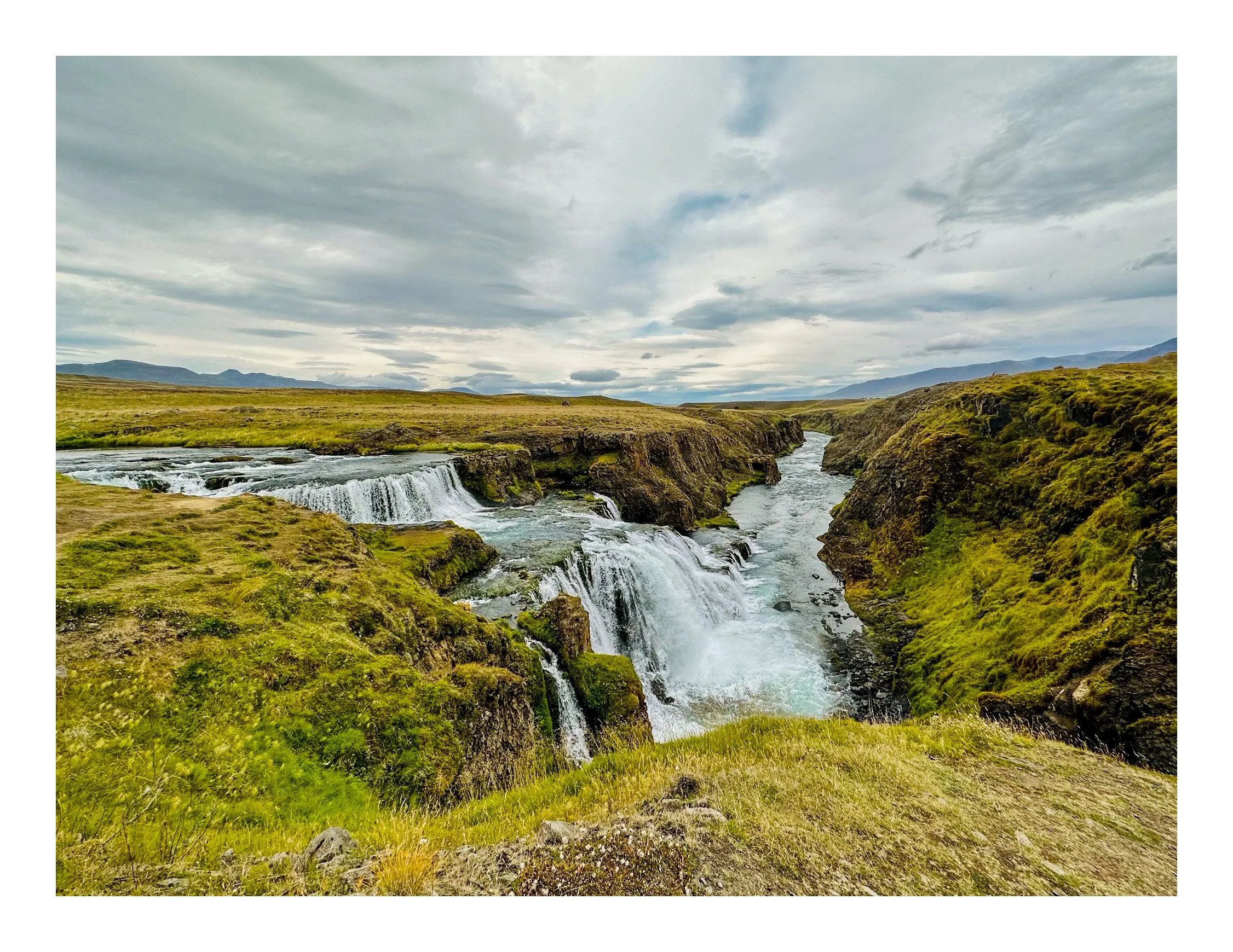 Scenic view of a waterfall cascading over rocky ledges into a river, surrounded by lush green moss-covered banks and hills under a cloudy sky.