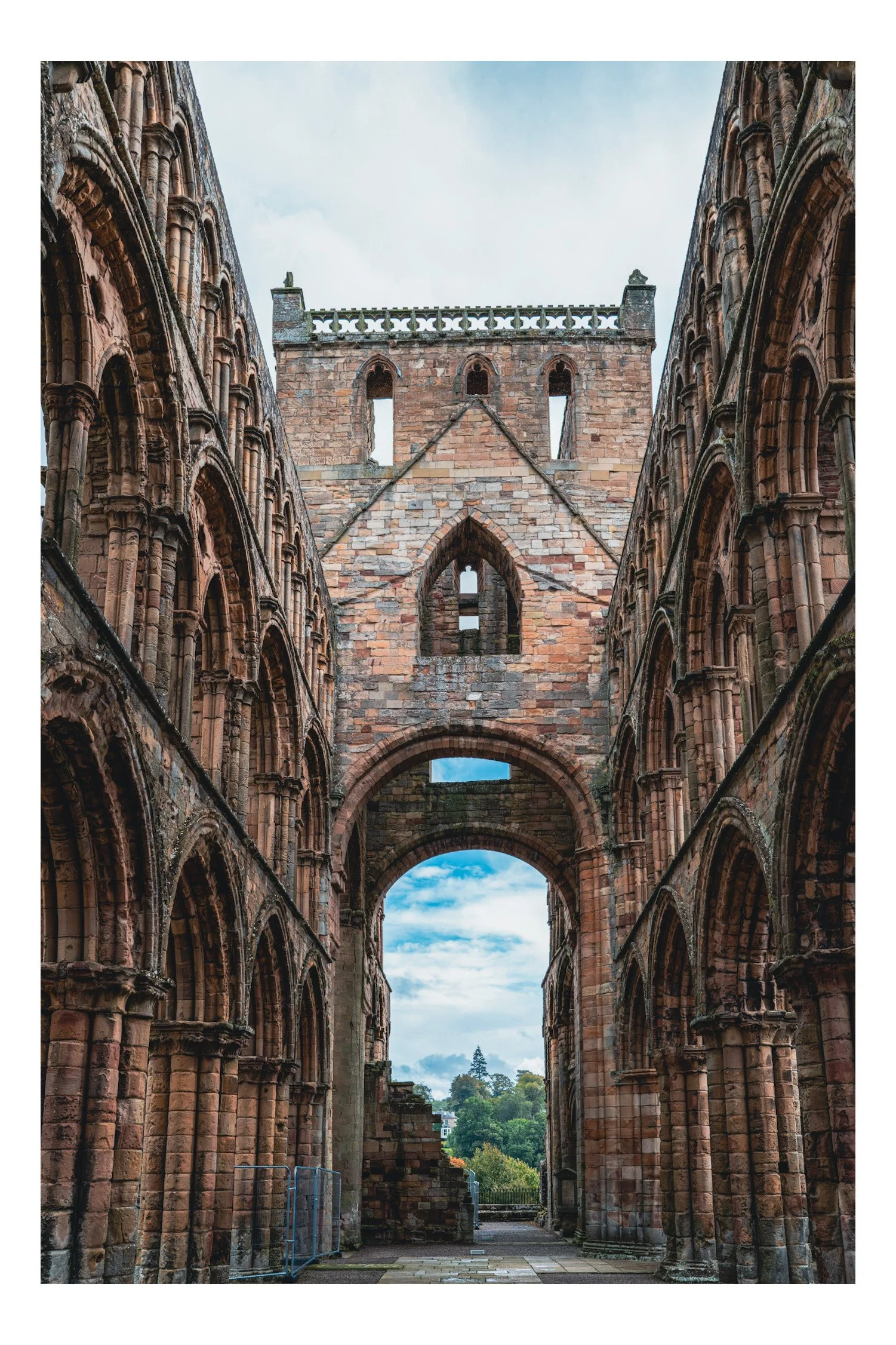 View of the ruins of an ancient stone cathedral with arches, windows, and a central arch opening to a cloudy sky and green landscape.