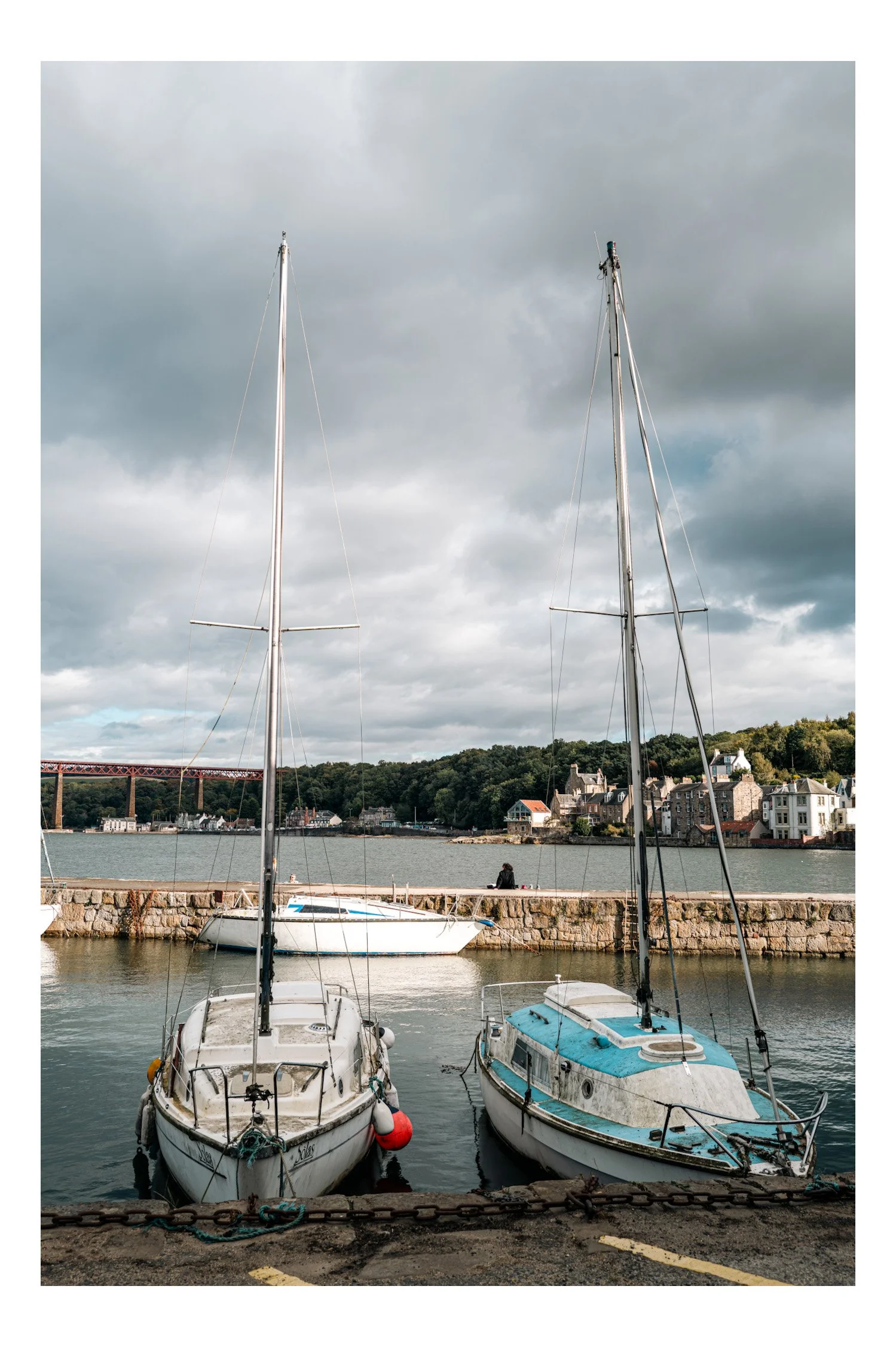 Two sailboats docked at a harbor with a stone seawall, with a person sitting on the seawall, and a coastal town with houses and greenery in the background under a cloudy sky.