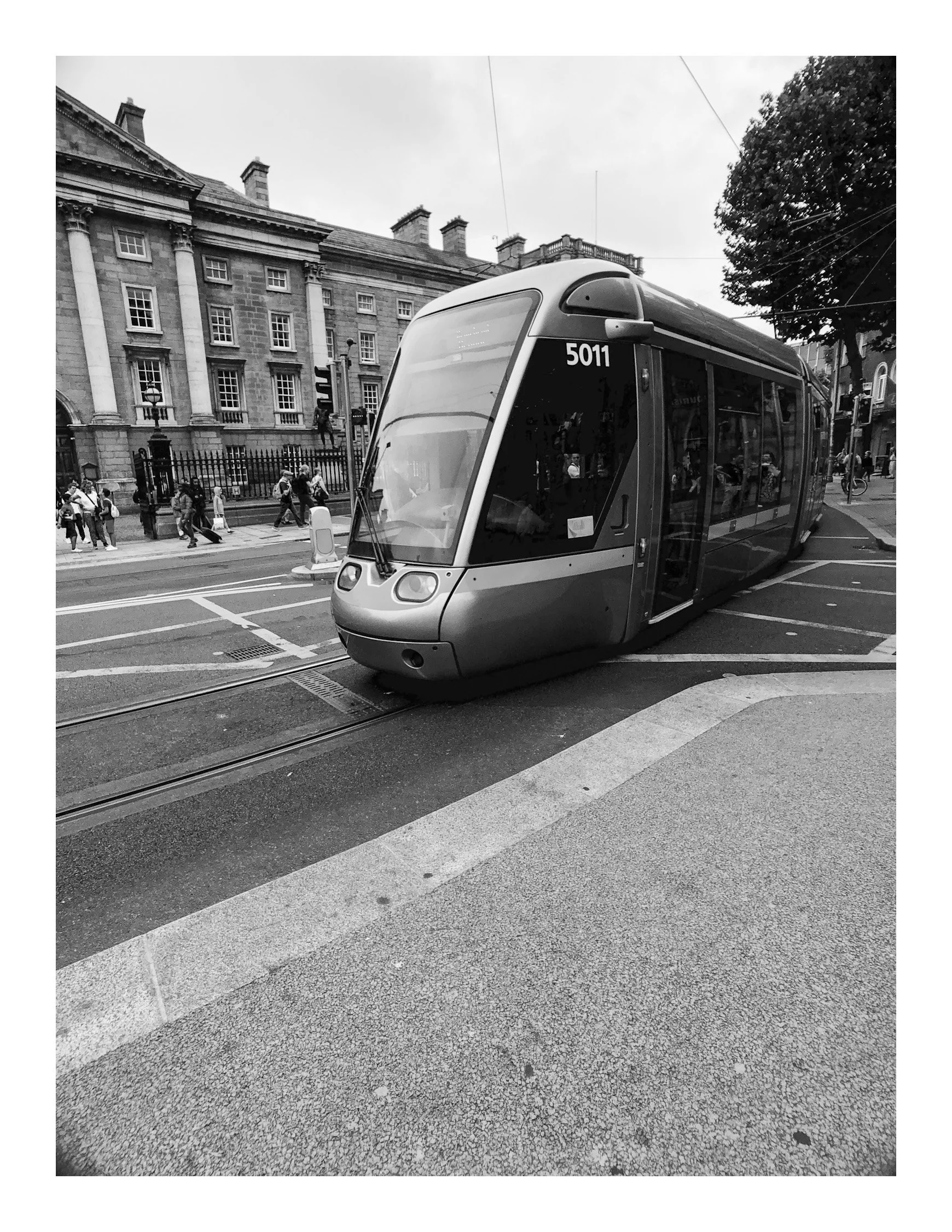 A modern tram with the number 5011 on the front, traveling along tracks on a city street, with pedestrians walking on the sidewalk and historic buildings in the background.