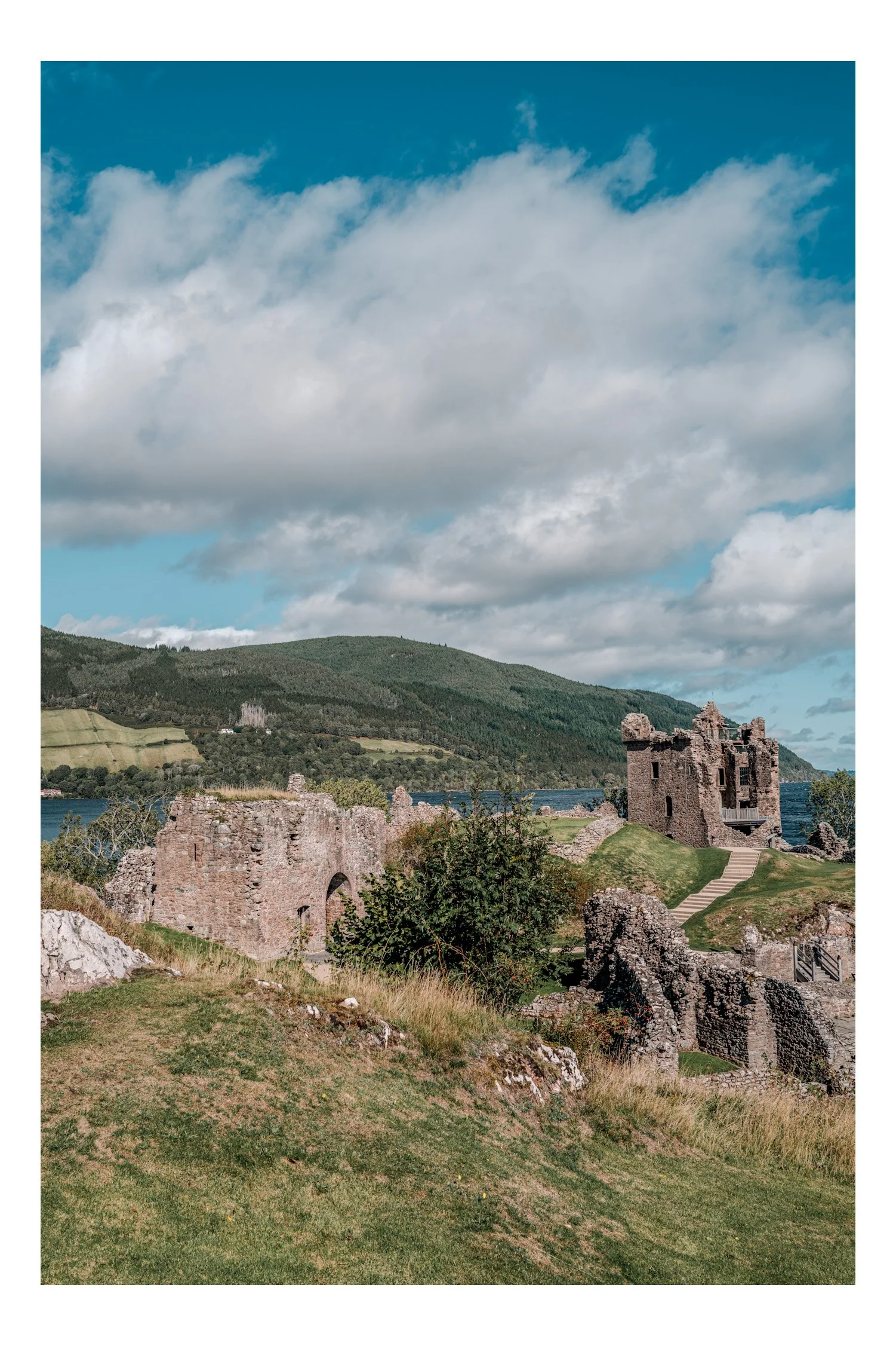Ruins of an old castle on a grassy hill with a lake and green hills in the background under a partly cloudy sky.