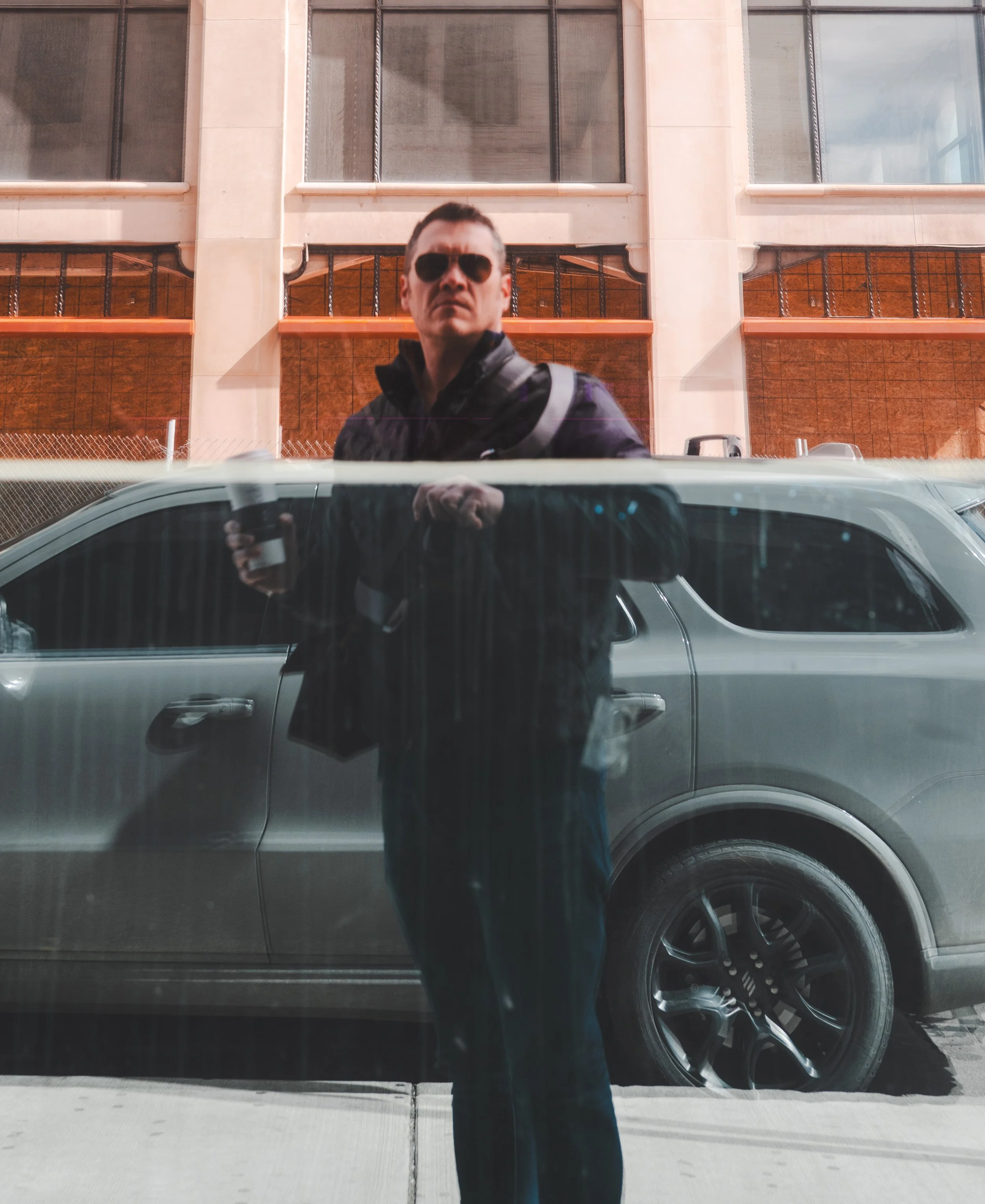 A man wearing sunglasses and a black jacket taking a photo of himself reflected in a glass window with a gray car parked behind him.