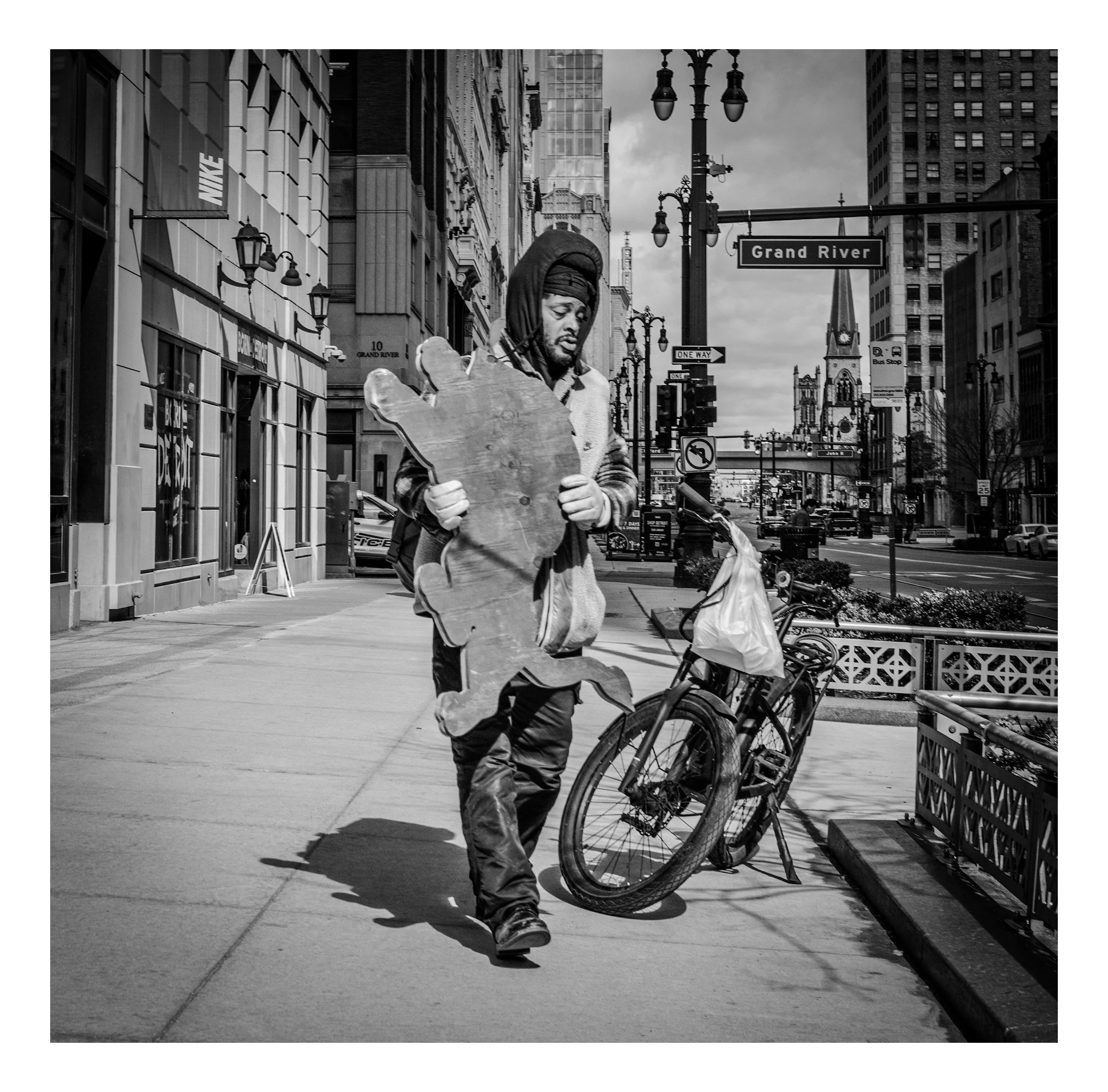 A man walking on city sidewalk holding a large, irregularly shaped wooden piece that resembles a map, with a bicycle parked nearby. The background shows tall buildings, street signs, and traffic lights, indicating an urban setting.