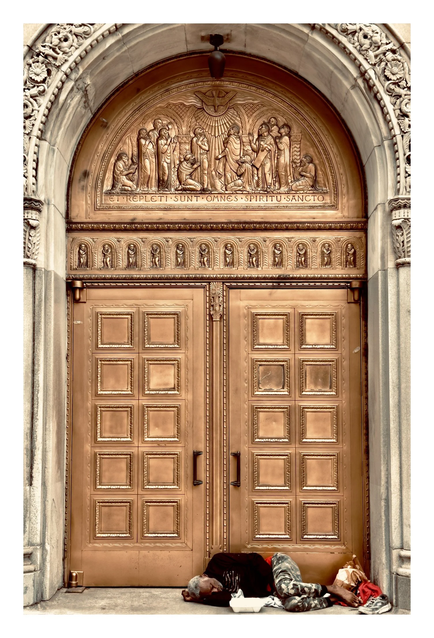 A large ornate cathedral door with detailed carvings and Latin inscription, with a man and a child sleeping on the ground in front of it.