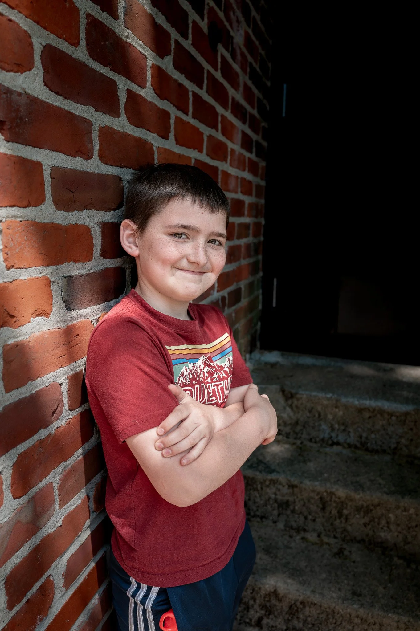 A boy with short dark hair, wearing a red graphic T-shirt and dark shorts, leaning against a red brick wall with arms crossed, smiling at the camera.