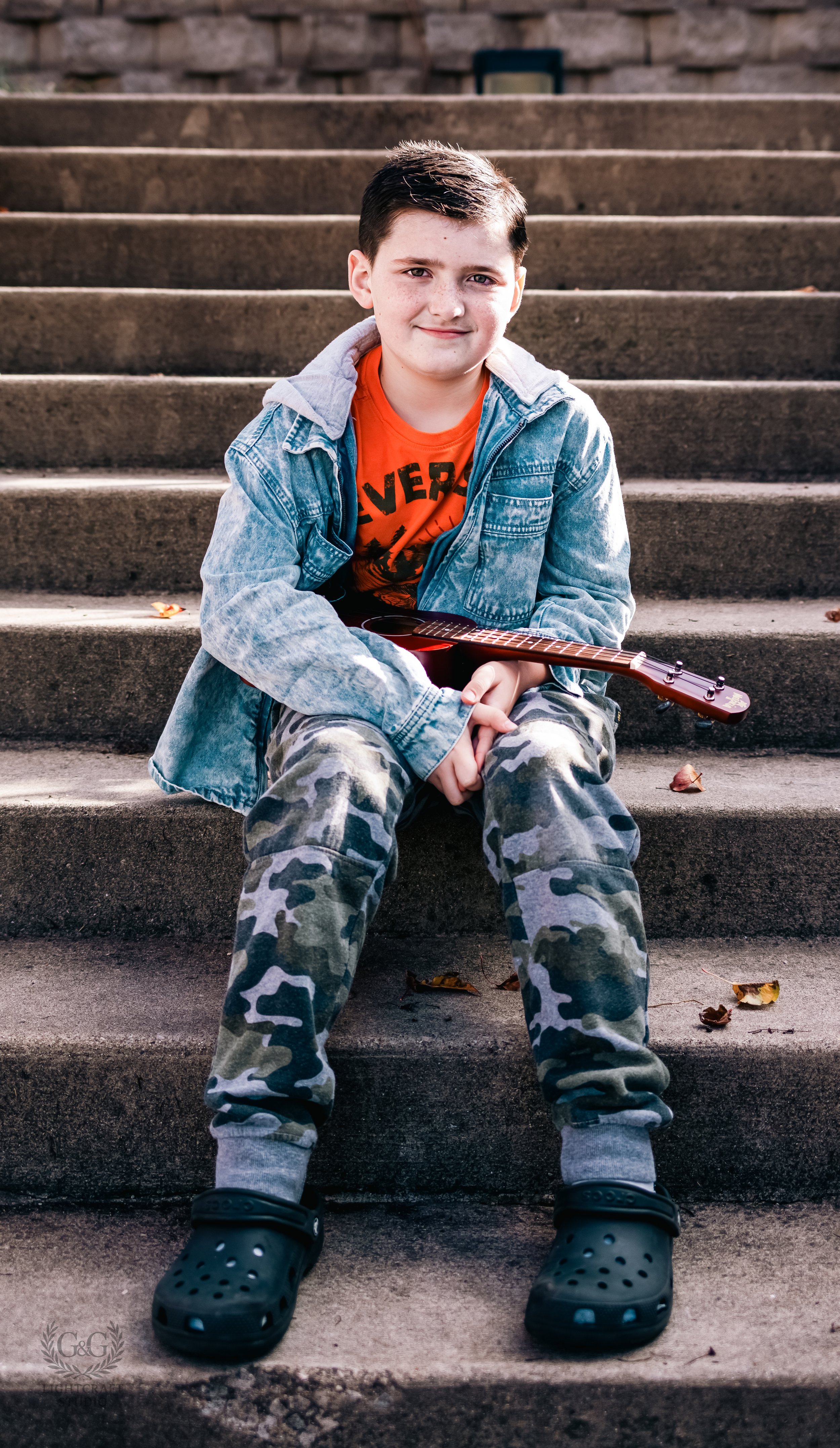 A boy with short dark hair, wearing a denim jacket, an orange T-shirt, camouflage pants, and black Crocs, sitting on outdoor concrete steps holding a small guitar.