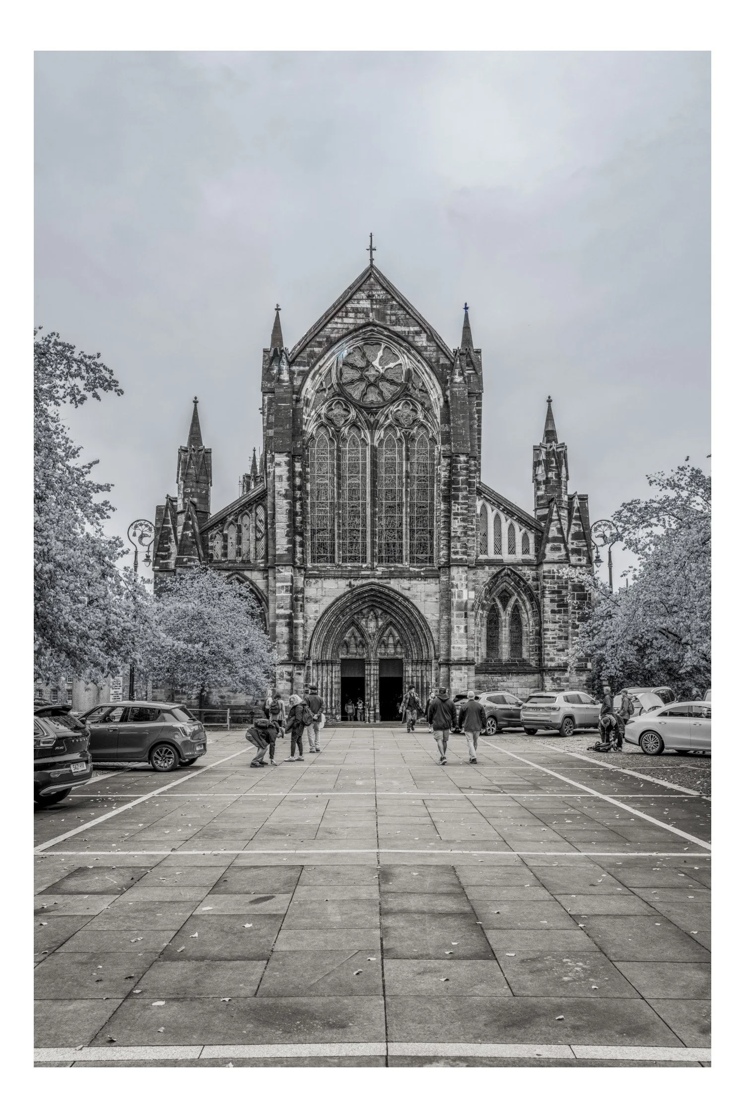 A large Gothic church with tall stained glass windows and pointed spires, viewed from the parking lot with several cars and people walking towards the entrance.