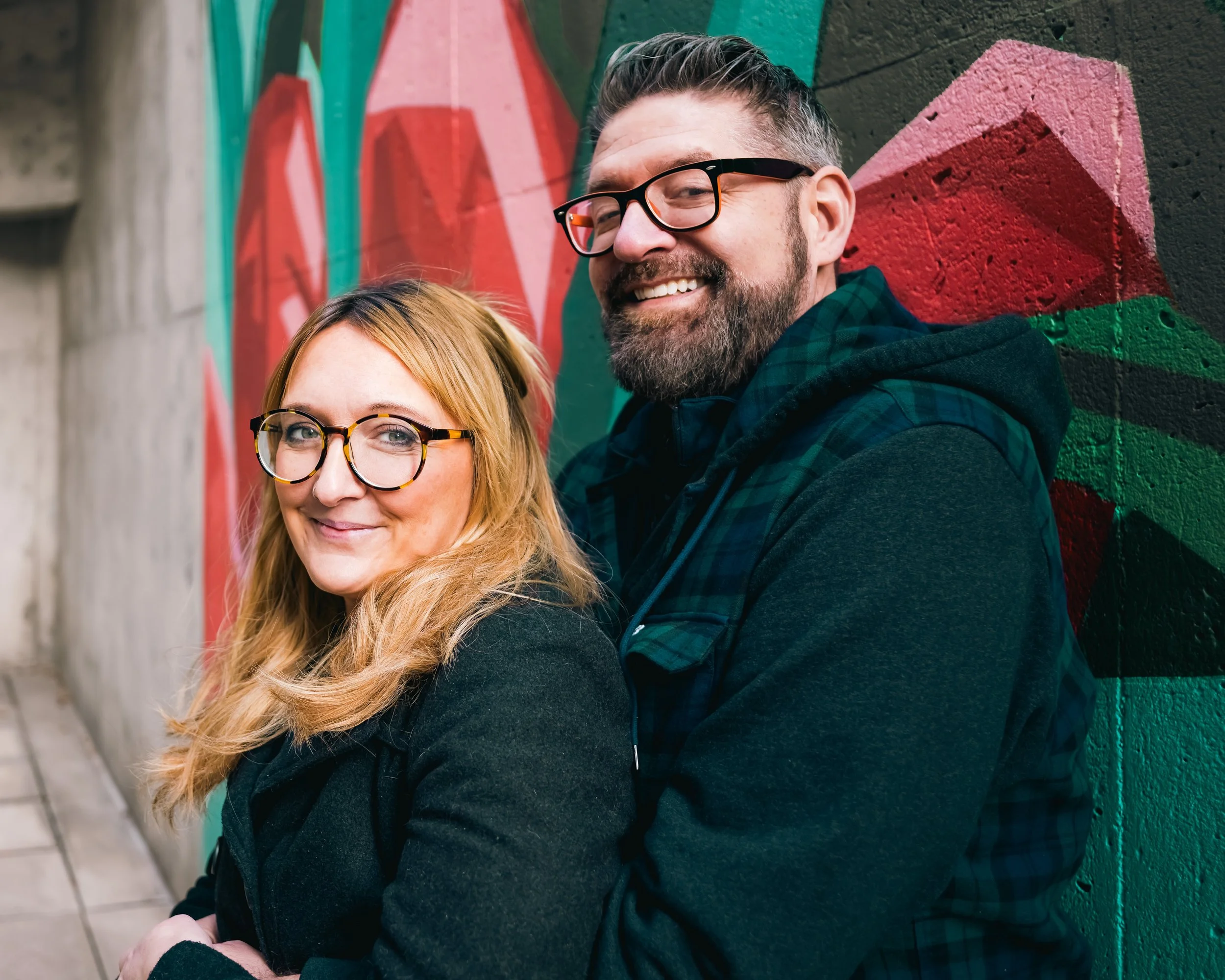 A smiling woman with glasses and long blonde hair and a smiling man with glasses and a beard standing close together against a colorful graffiti wall.