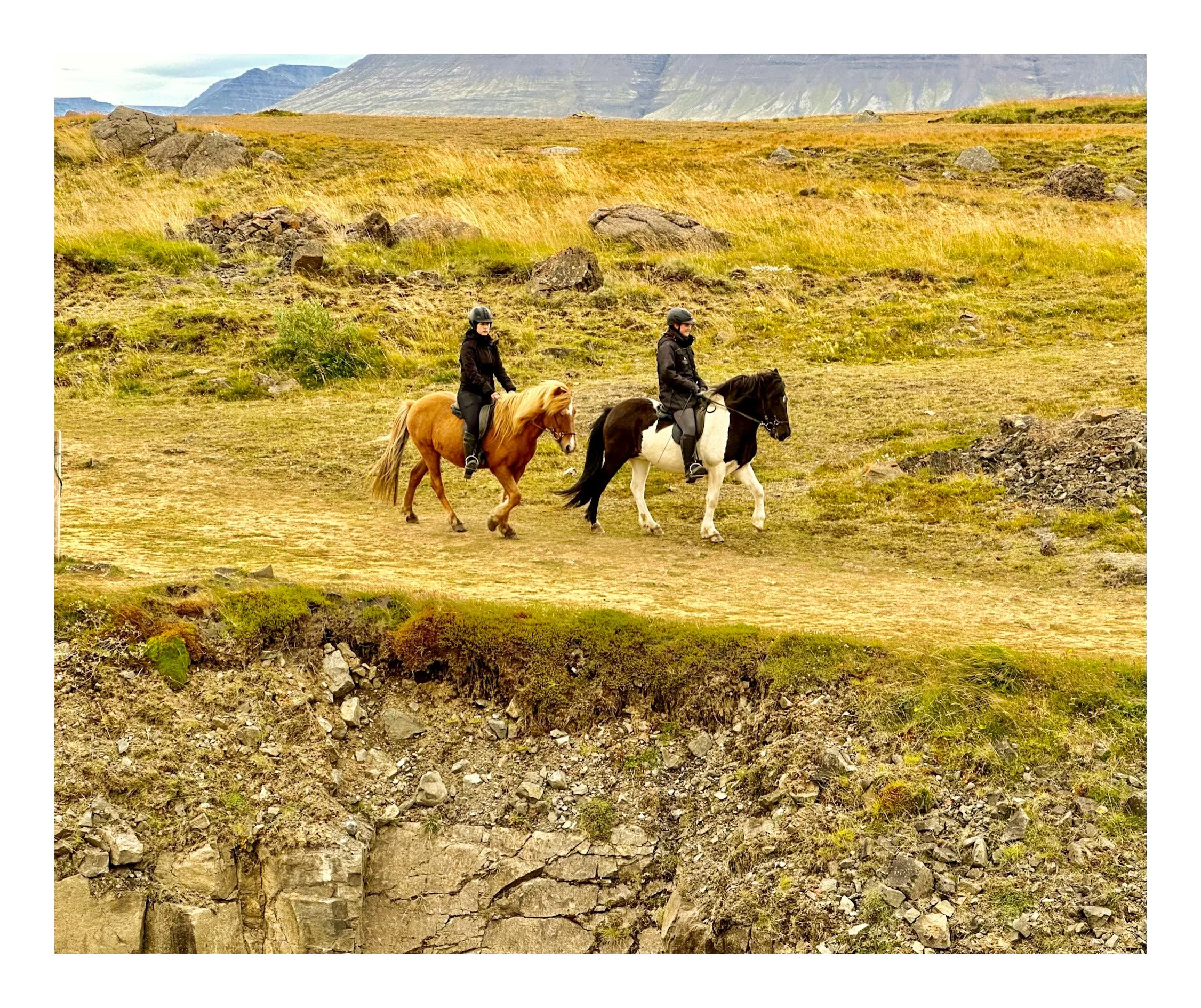 Two people riding horses on a dirt trail in a grassy landscape with mountains in the background.