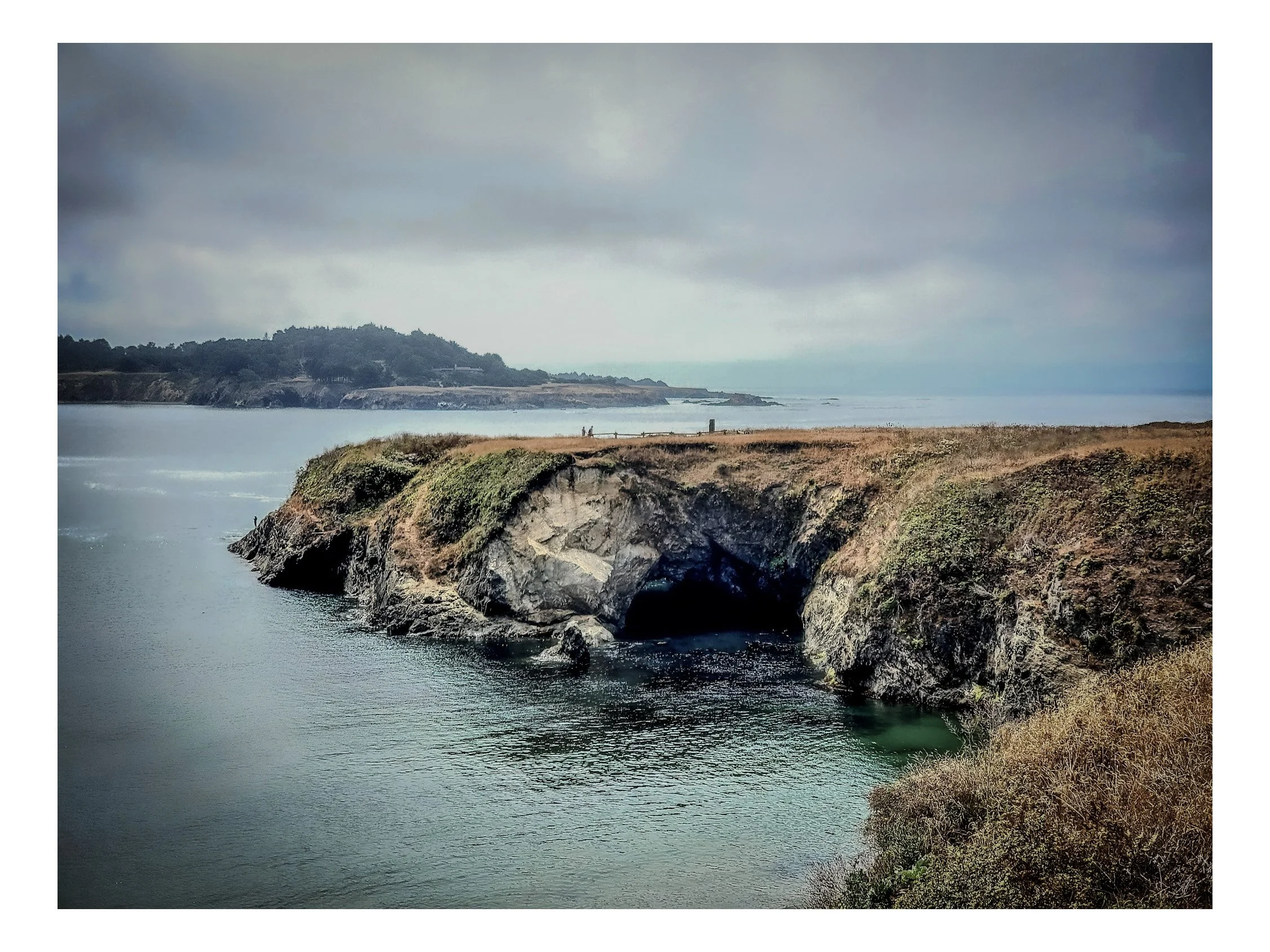 Coastal landscape with rocky cliffs, water below, grassy and shrub-covered land, cloudy sky, and distant hills.