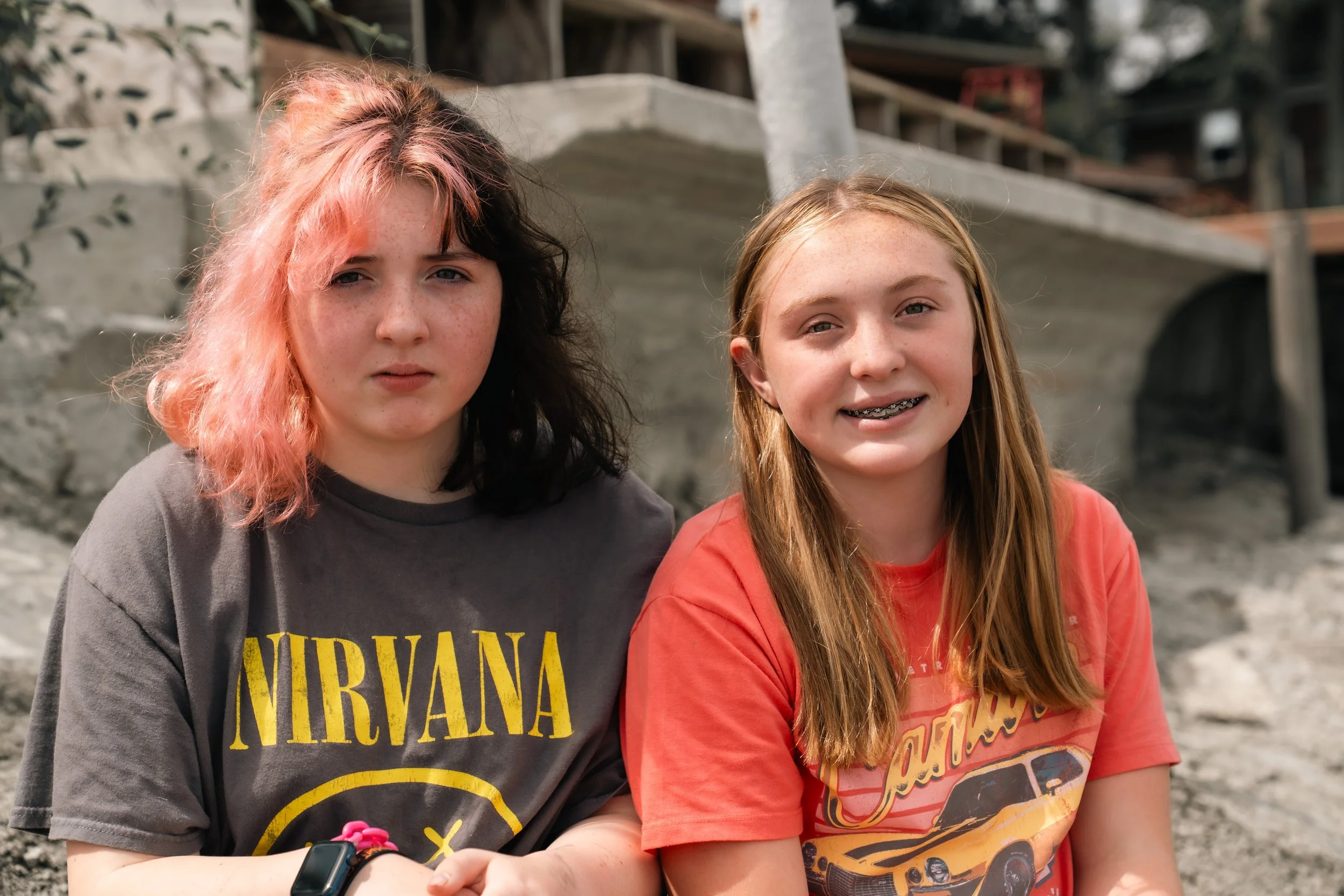 Two young girls sitting outdoors. The girl on the left has pink and black hair, wearing a Nirvana T-shirt and a wristwatch. The girl on the right has long blonde hair, braces, and is wearing a red T-shirt with a car graphic. They are sitting on a roc