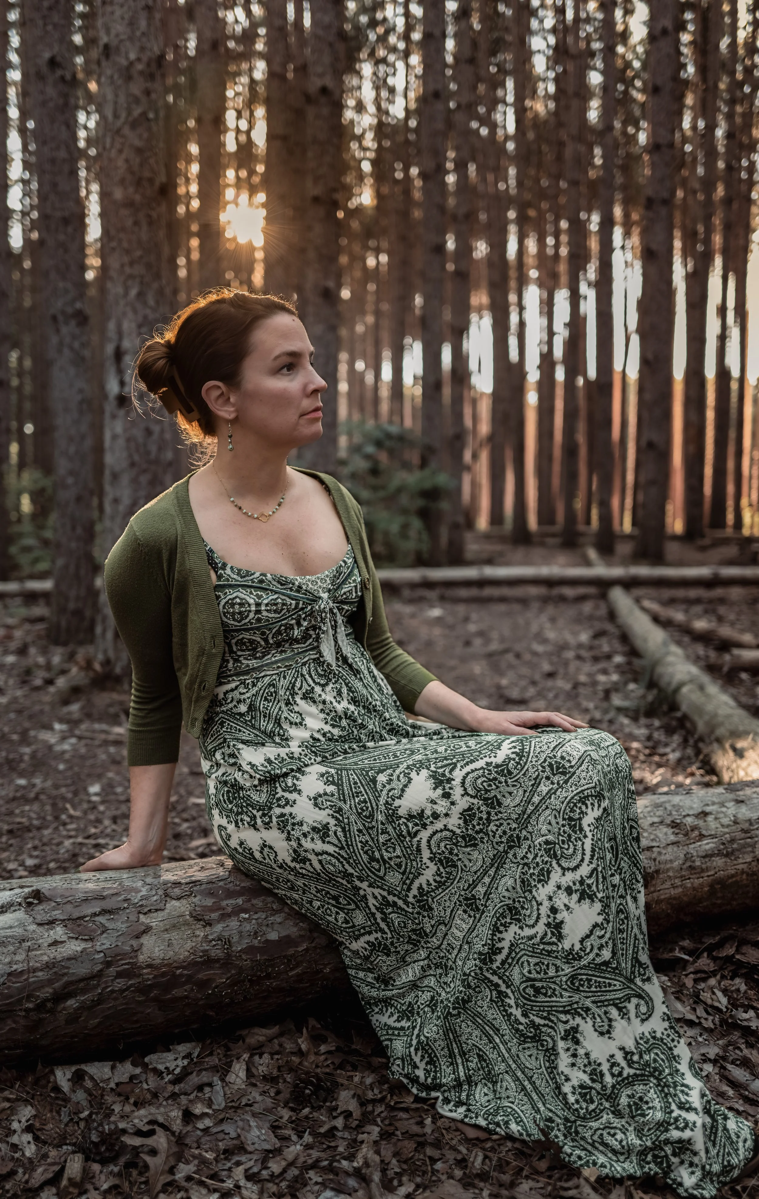 A woman sits on a fallen log in a forest during sunset, wearing a patterned dress and a green cardigan, with trees and sunlight in the background.