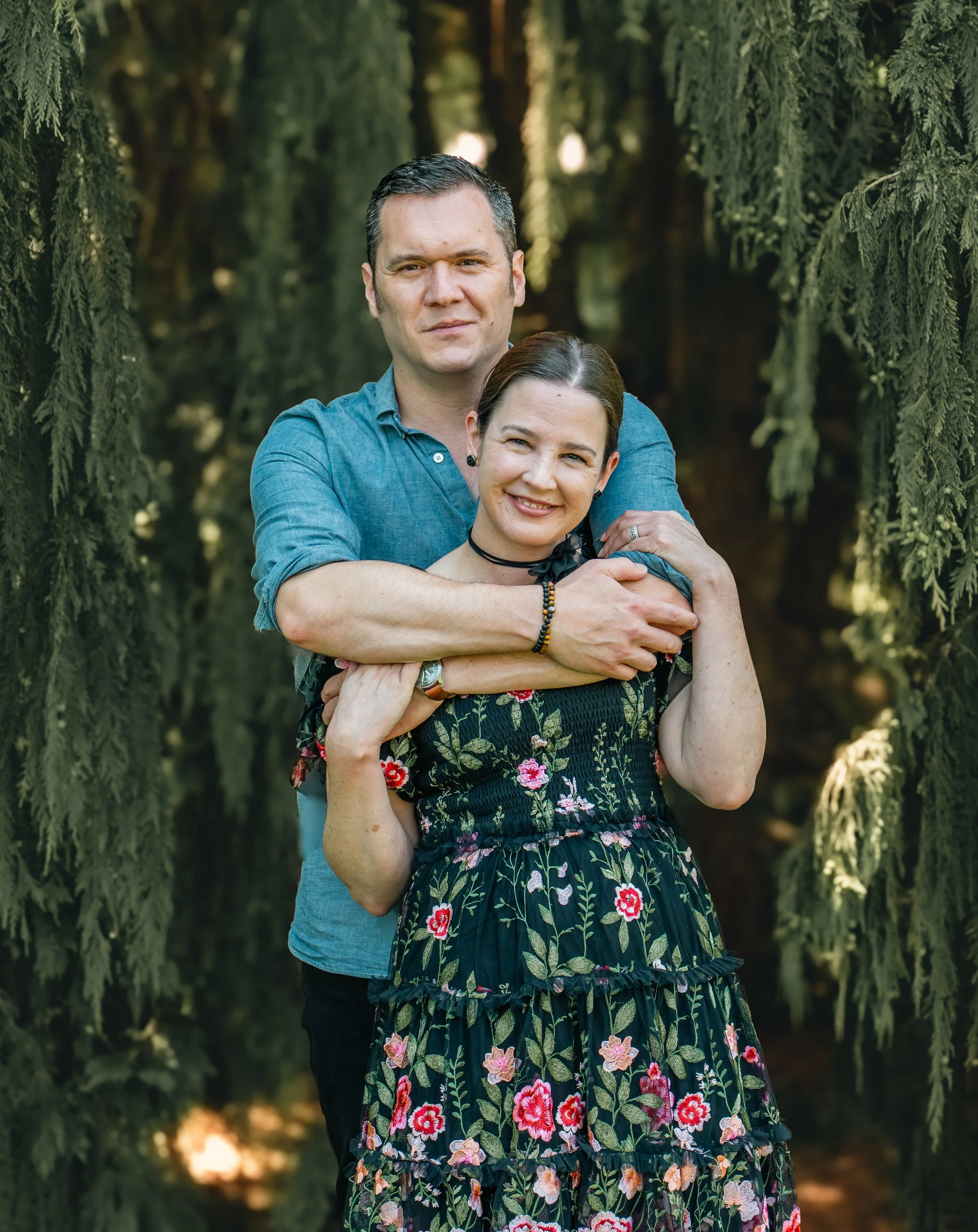 A couple stands outdoors among trees, with the man embracing the woman from behind. The woman smiles at the camera, wearing a floral dress, with dark hair pulled back, and the man has short dark hair, wearing a blue shirt.