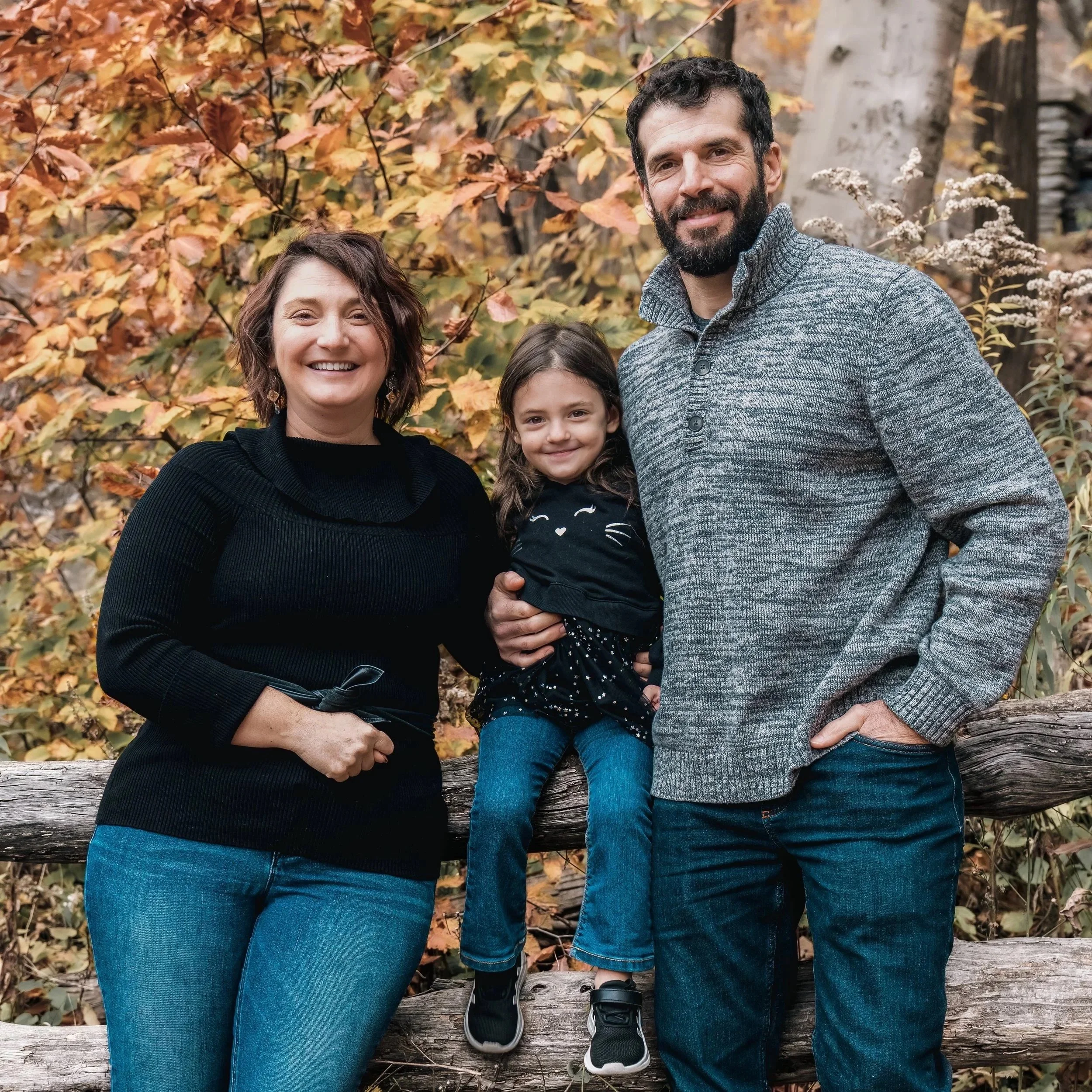 A family of three standing outdoors during autumn, surrounded by colorful fall leaves, smiling at the camera.
