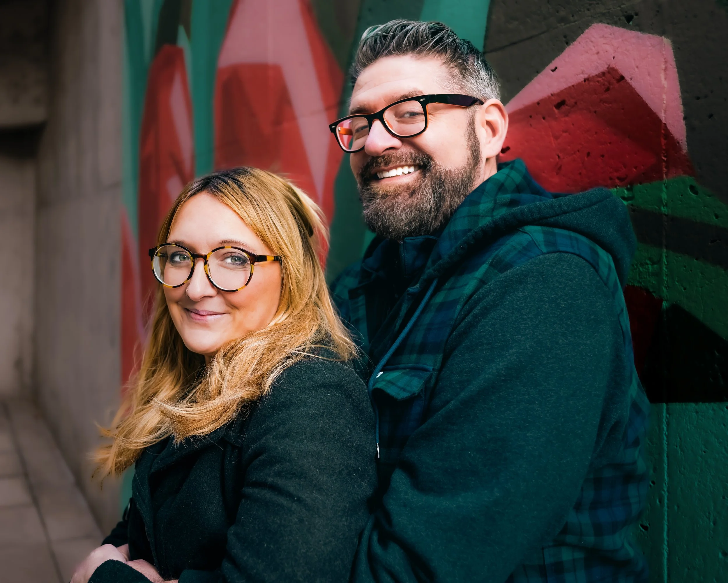 A smiling man and woman with glasses posing outdoors against a colorful graffiti wall.