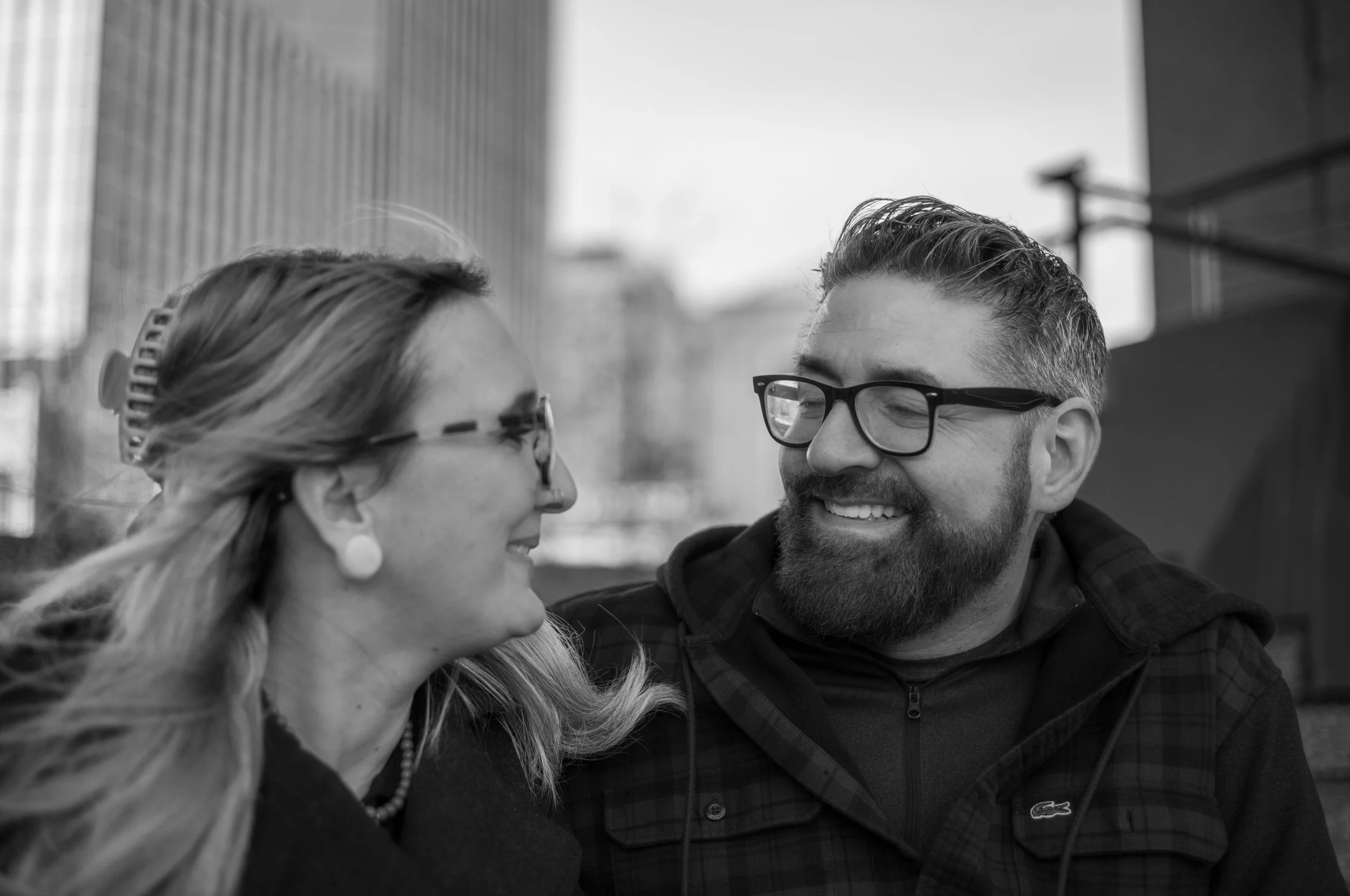 A black-and-white photo of a smiling man and woman looking at each other outdoors in an urban setting.