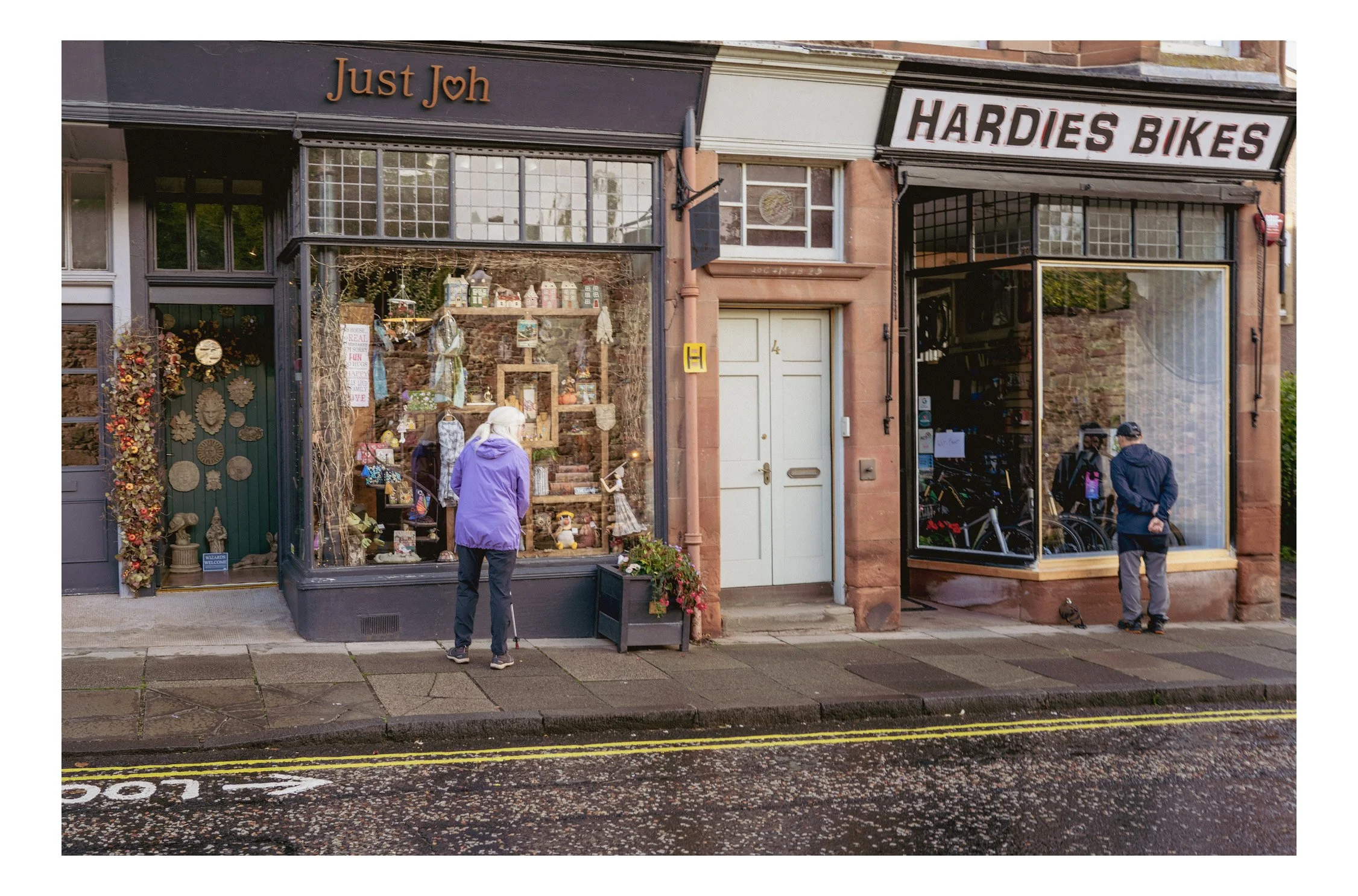 Street view of two shops, 'Just Joh' on the left with a window display of decorative items and 'Hardies Bikes' on the right with bike inside, two people looking into shop windows, wet pavement, and cloudy weather.