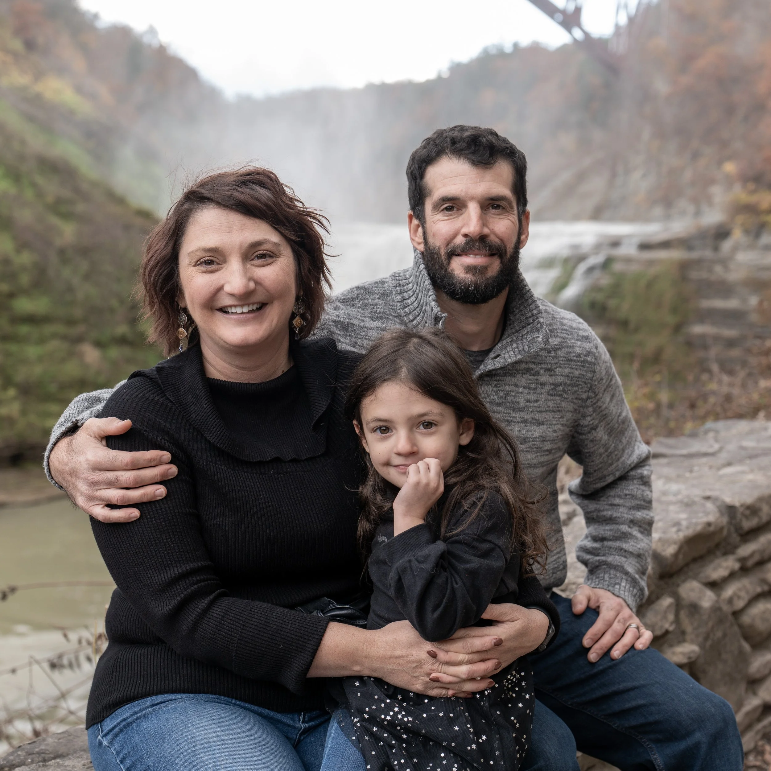 A family of three with a waterfall in the background, sitting outdoors on a rocky ledge.