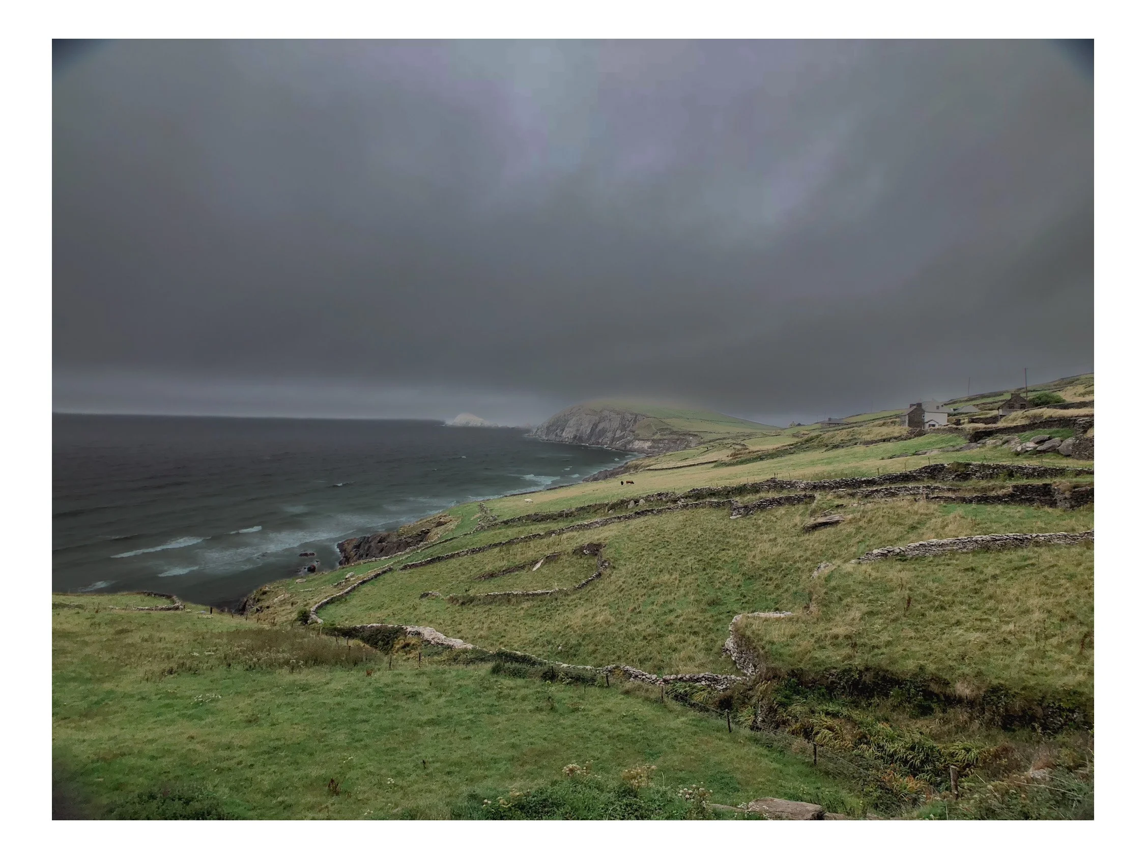 Coastal landscape with green rolling hills, stone walls, a few scattered houses, and a dark cloudy sky over the ocean.