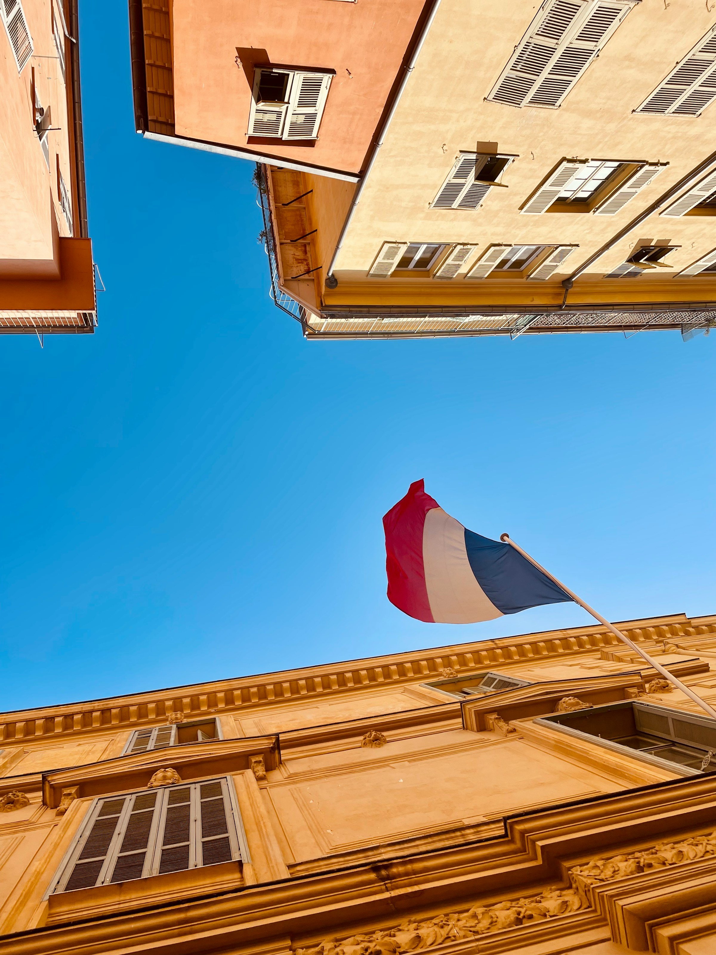 Looking up at a clear blue sky, framed by buildings with closed shutters, and a flagpole with a waving French flag.