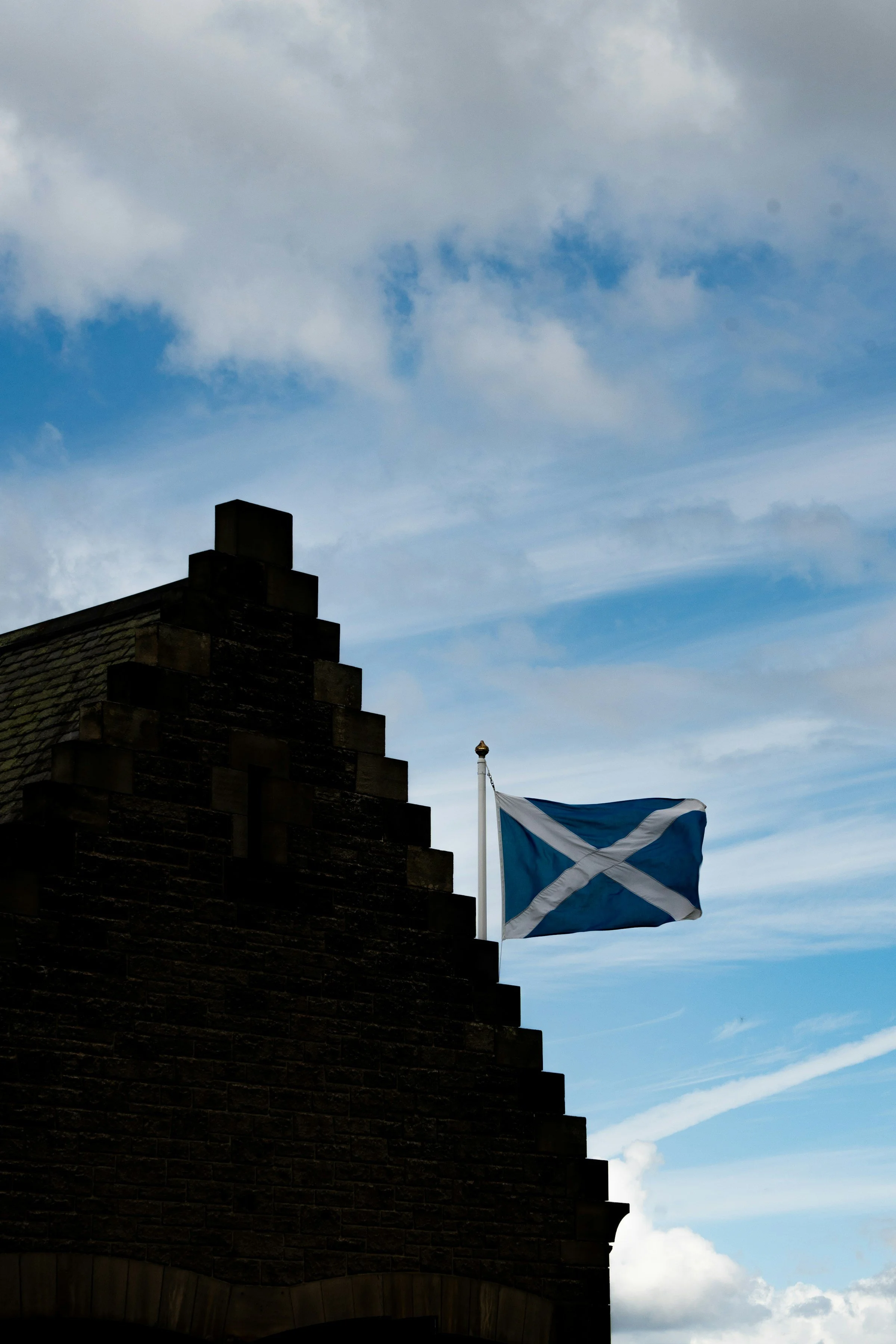 A Scottish flag flying on a pole against a blue sky with clouds, atop a stone building with a stepped design.
