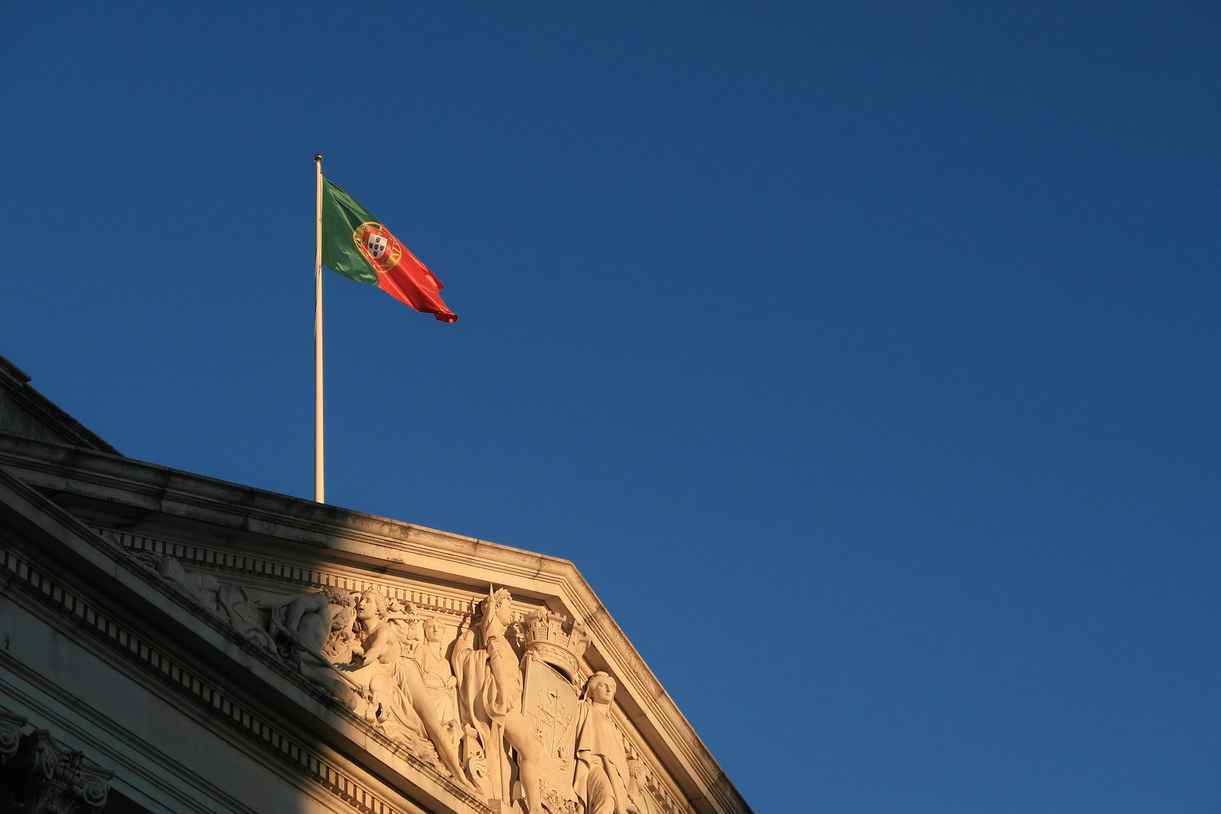Portuguese flag flying above a classical building with detailed sculptures, against a clear blue sky.