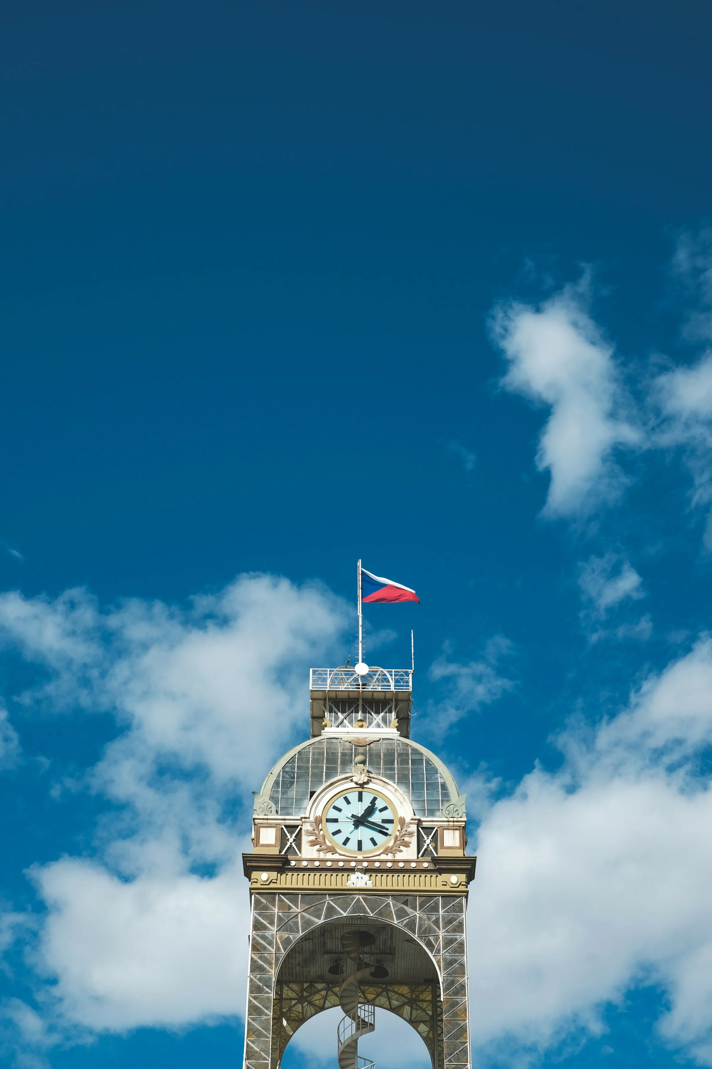 A clock tower with a clock showing the time as 1:25, topped with a flagpole flying the Czech flag, set against a partly cloudy blue sky.