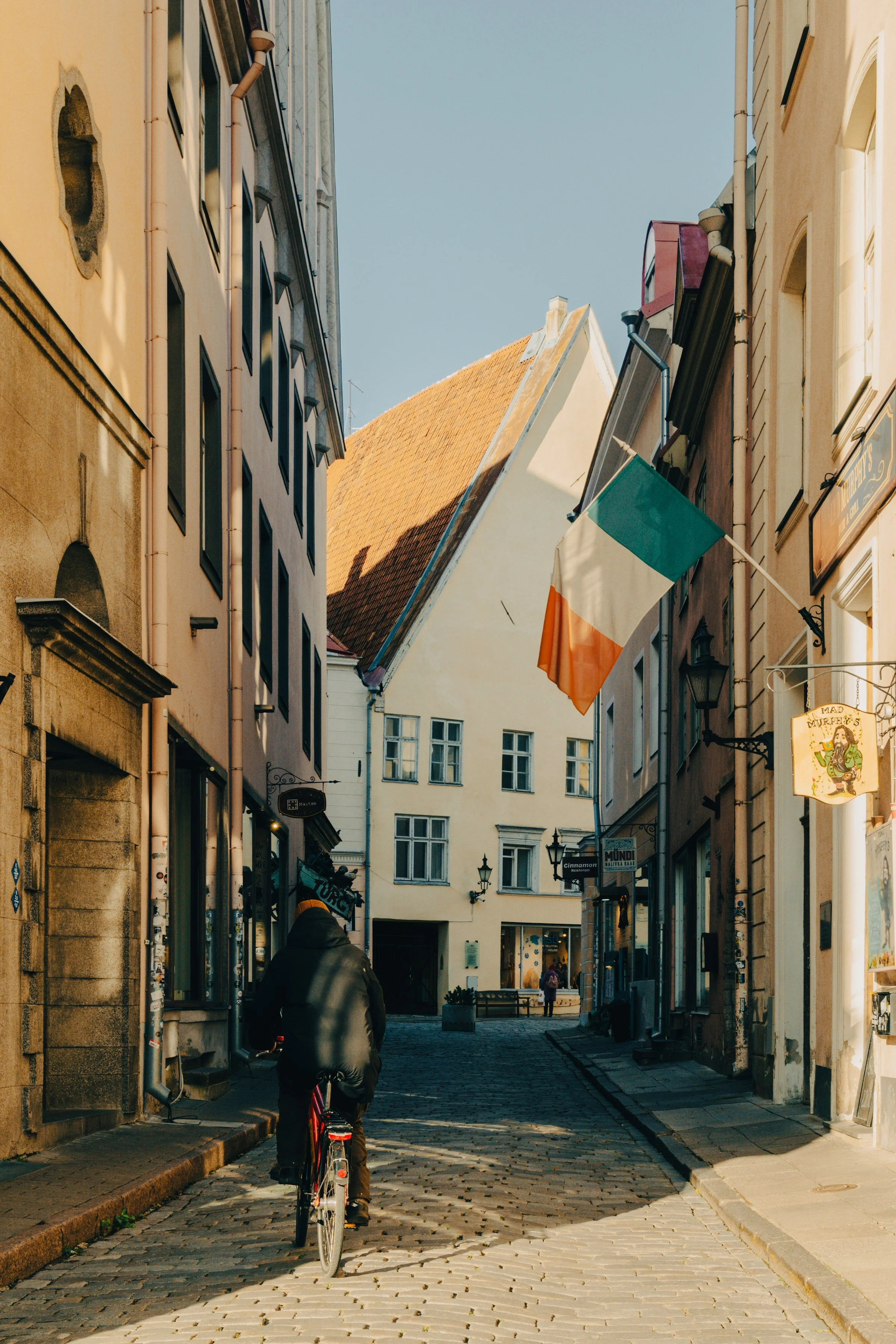 A person riding a bicycle down a narrow, cobblestone street lined with buildings in an European town. An Irish flag hangs from a building on the right side of the street. The weather is sunny with a clear sky.