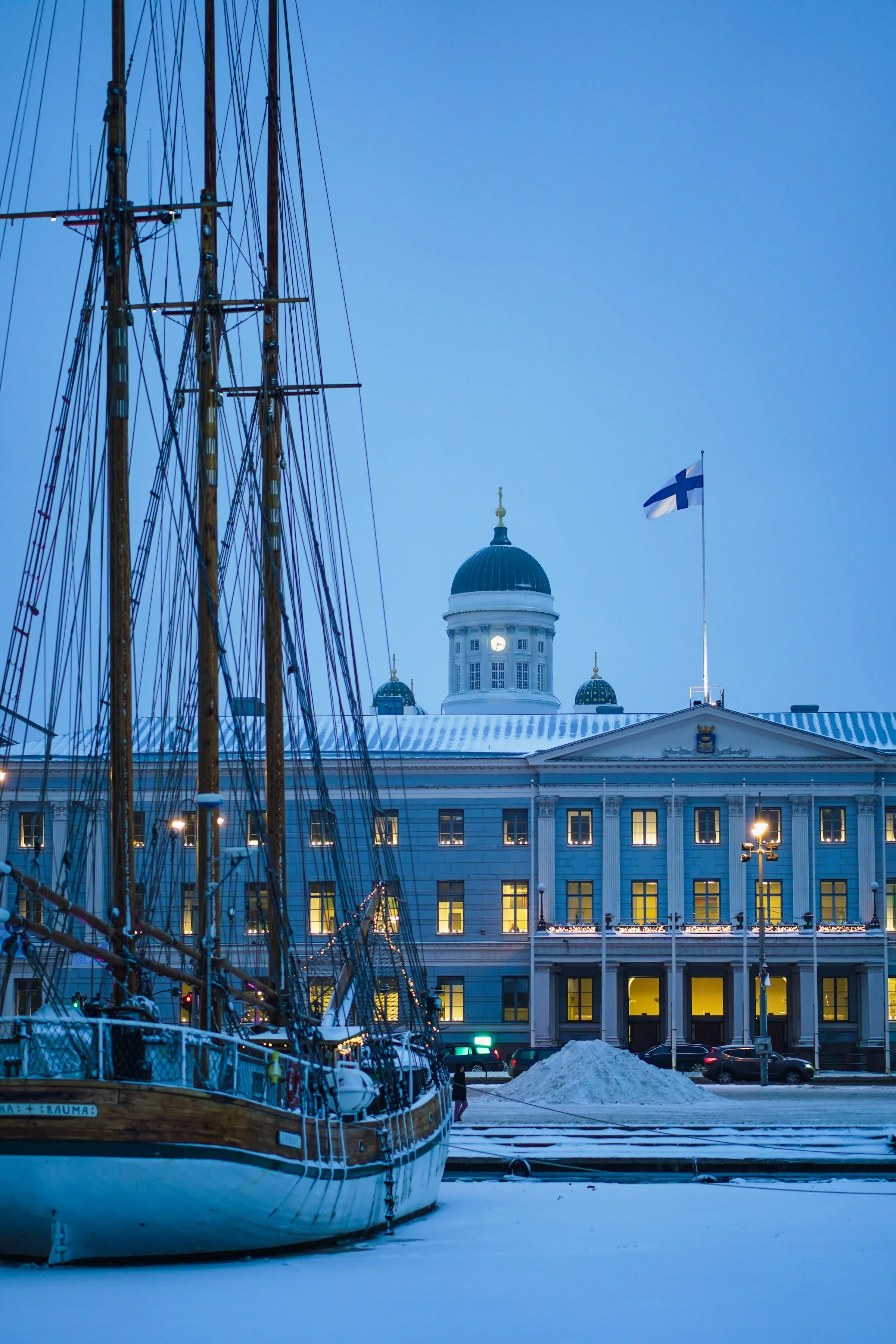 A historic building with a dome and Finnish flag, a sailing ship with masts, and snow-covered ground in Helsinki, Finland.