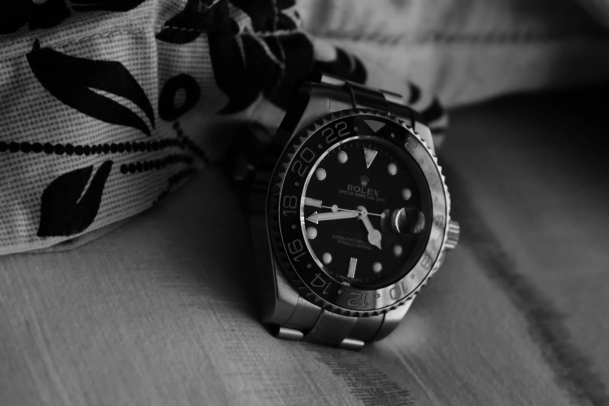 A men's wristwatch with a black dial, metal bezel, and metal bracelet, resting on a fabric surface with a patterned cloth nearby.