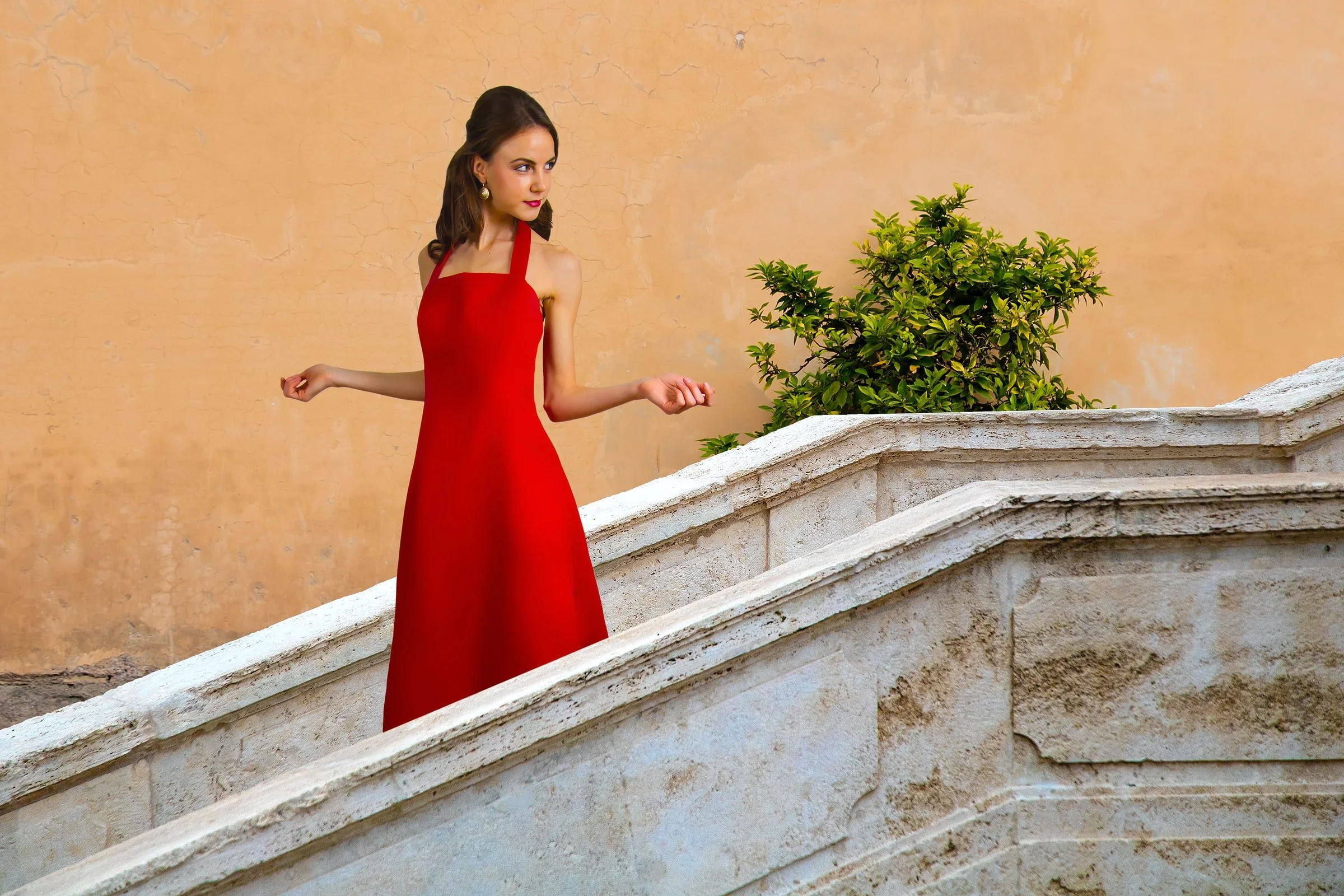 A woman with brunette hair, wearing a floor-length red dress with spaghetti straps, standing on a stone staircase outdoors next to a green bush against a textured beige wall.