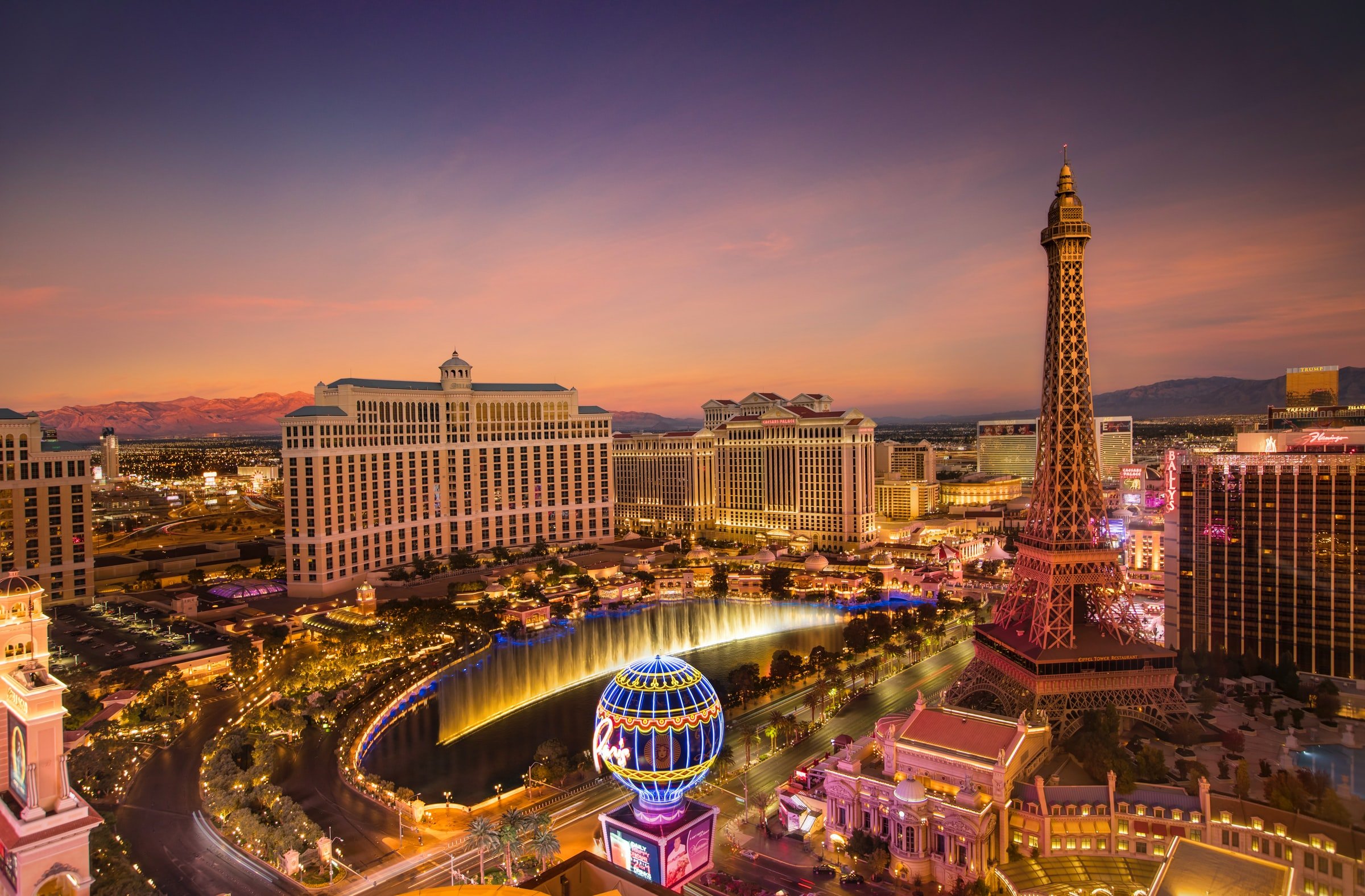 Las Vegas Strip cityscape at sunset featuring the Paris Hotel replica of Eiffel Tower, Bellagio fountains, and various hotels and casino buildings with colorful lights.