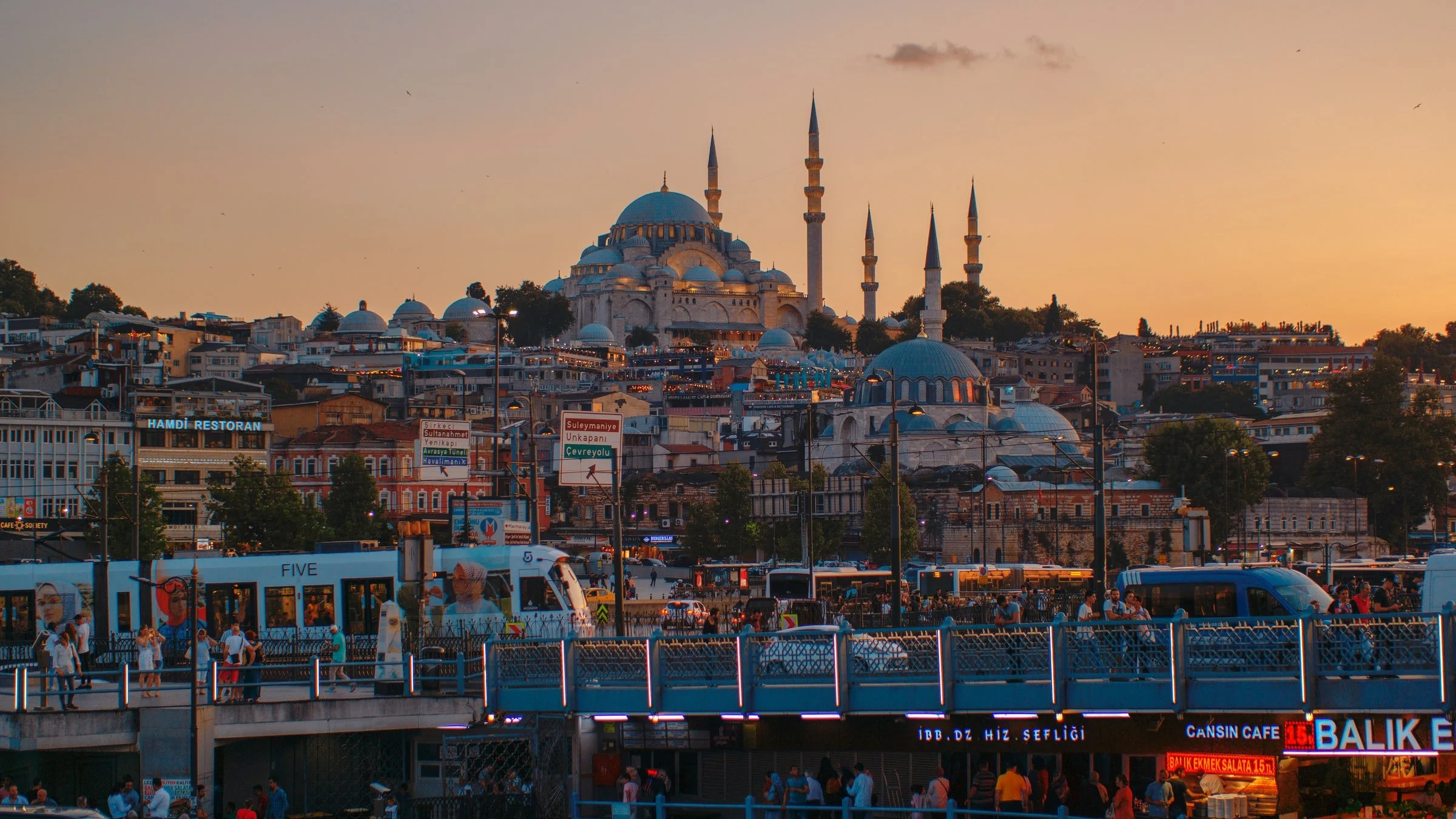 Sunset view of Istanbul with the Suleymaniye Mosque on a hill in the background, bustling city streets and activities in the foreground, including pedestrians, vehicles, and signs.