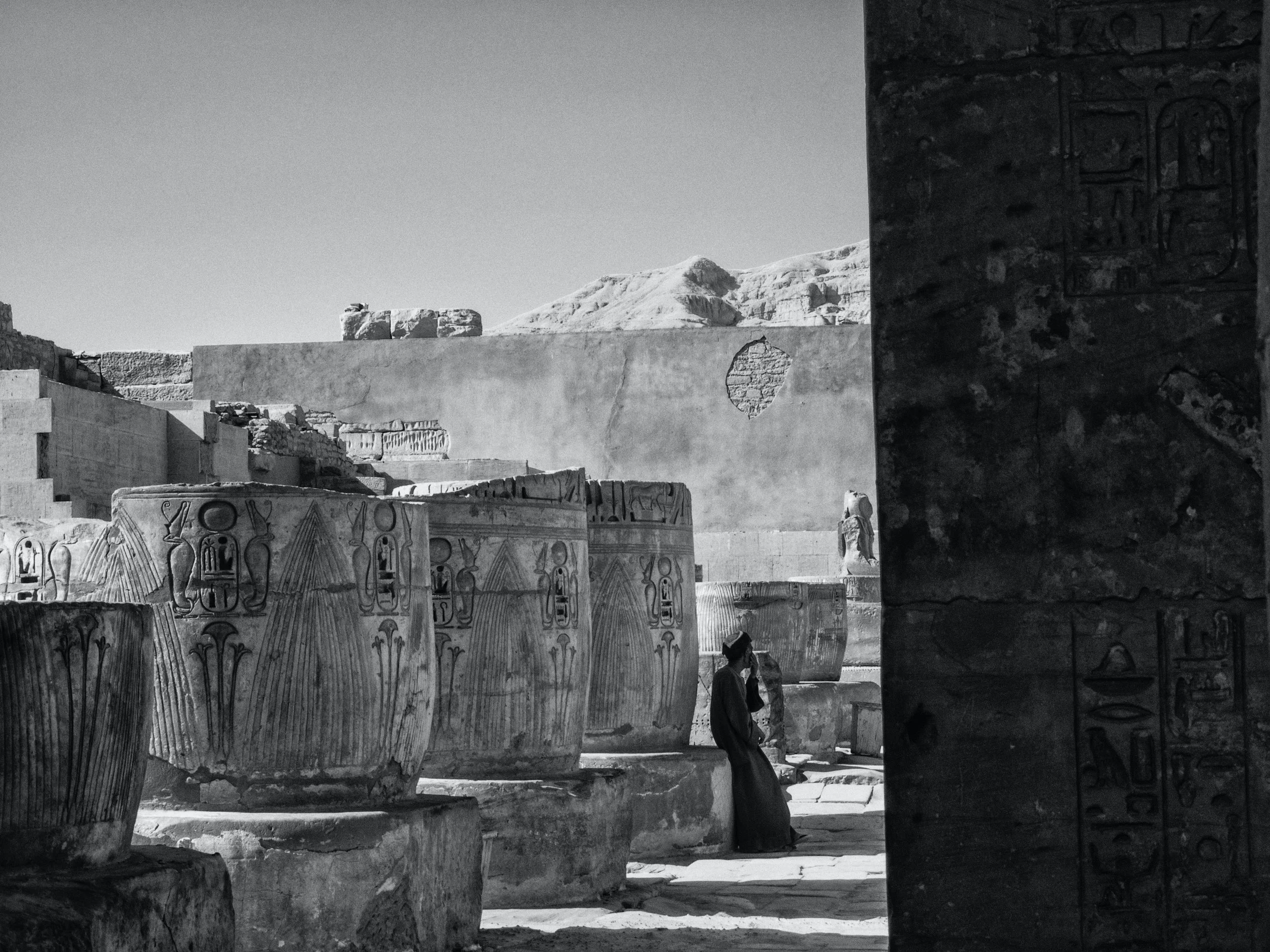 A person in traditional clothing standing among ancient Egyptian ruins with hieroglyphs on stone columns and a mountain in the background.