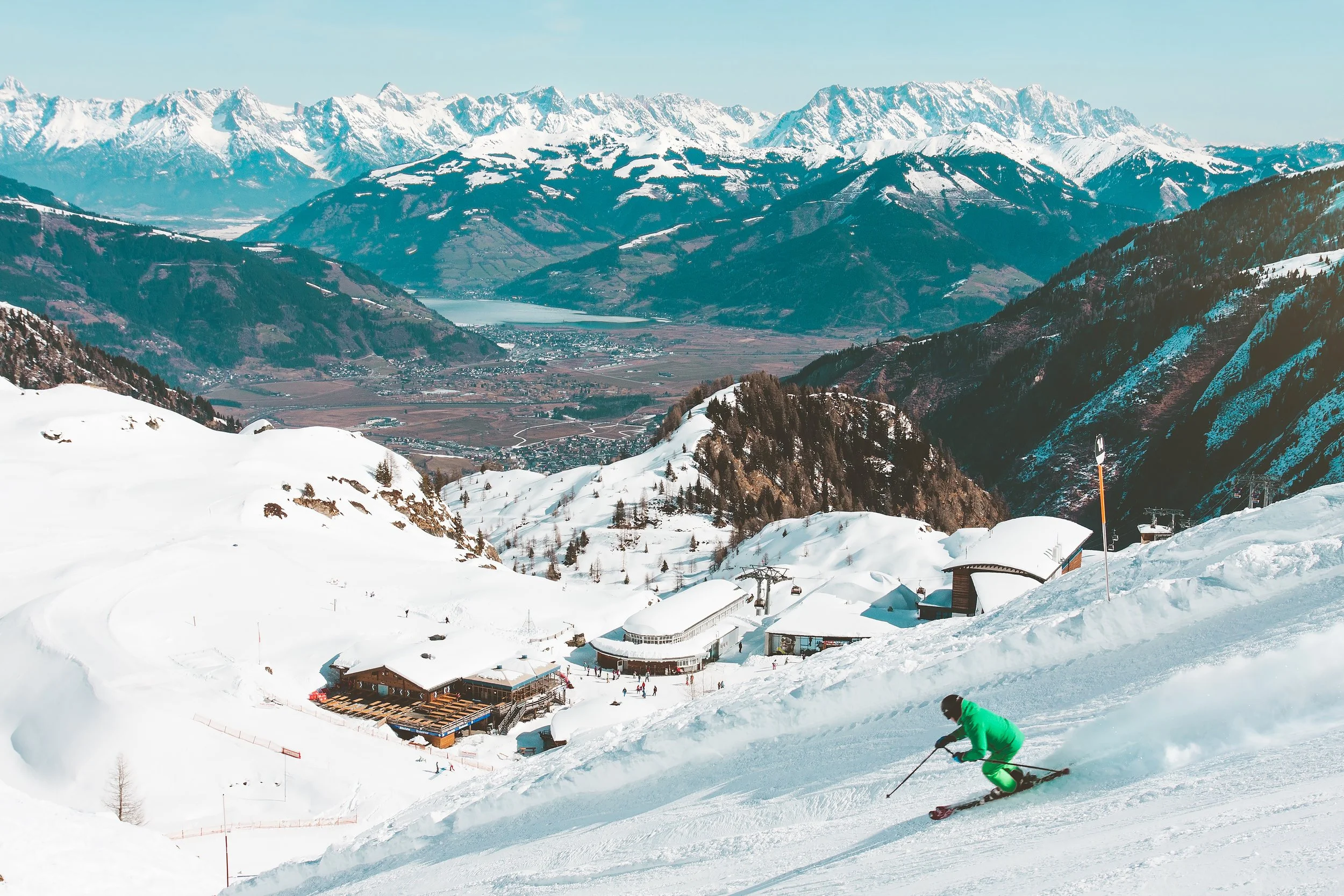 A person in a green ski suit skiing down a snowy mountain slope with a mountain lodge in the background, overlooking a valley with a lake and snow-capped peaks.