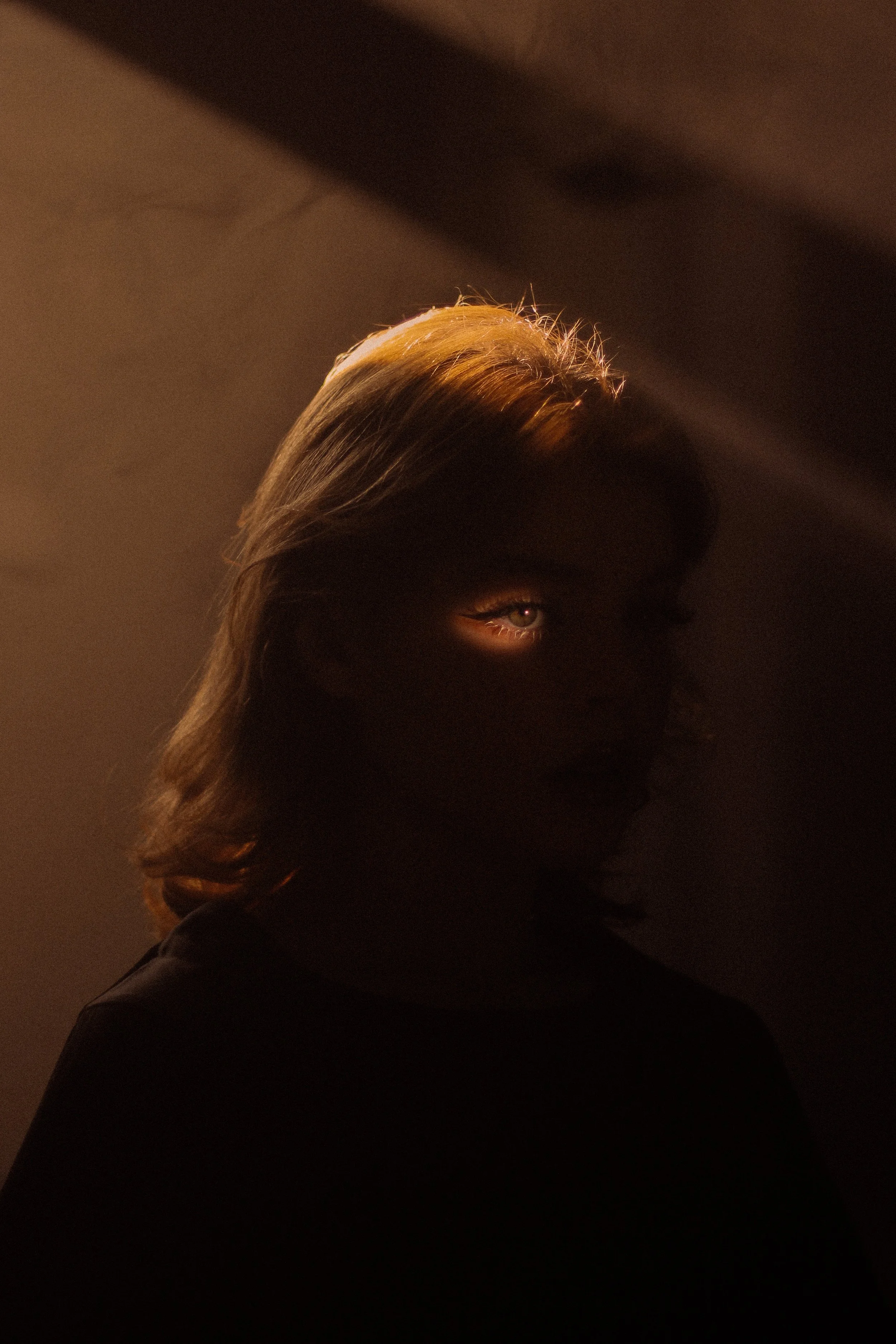 A woman with shoulder-length hair illuminated by a light that partially reveals her face and one eye, with the rest of her face mostly in shadow, against a dark background.