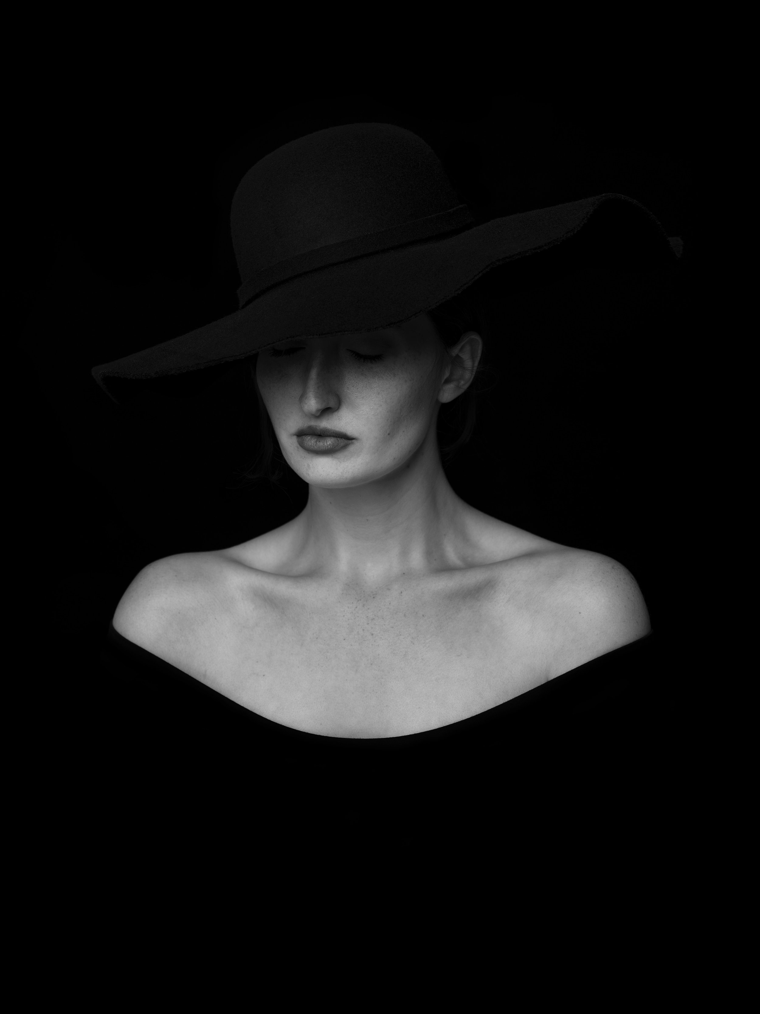 Black and white portrait of a woman with fair skin, wearing a large wide-brimmed hat that covers part of her face, with her eyes closed and head slightly tilted down.
