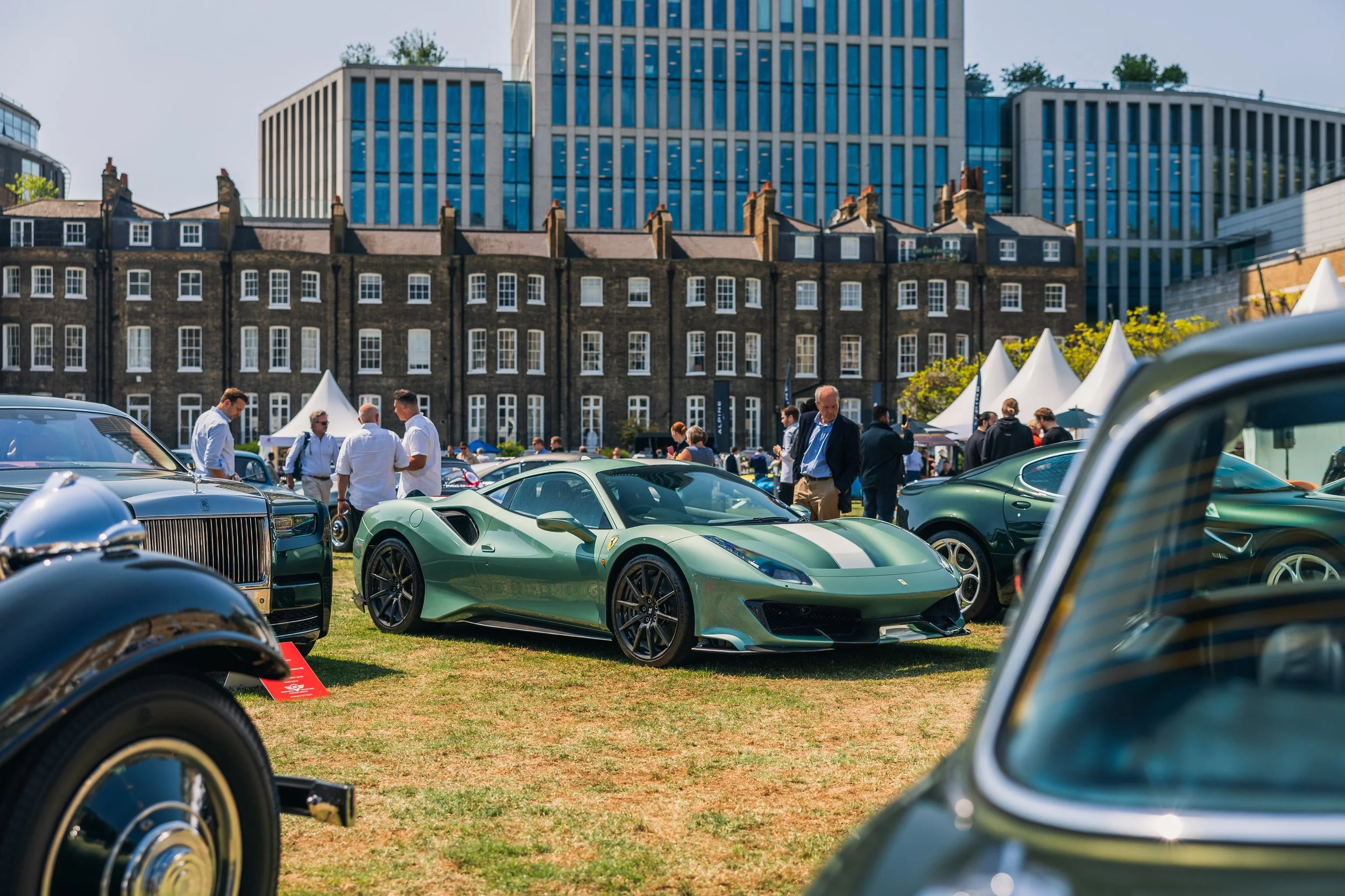 Green Ferrari sports car on display at a car show outdoor event with people viewing cars and modern buildings in the background.