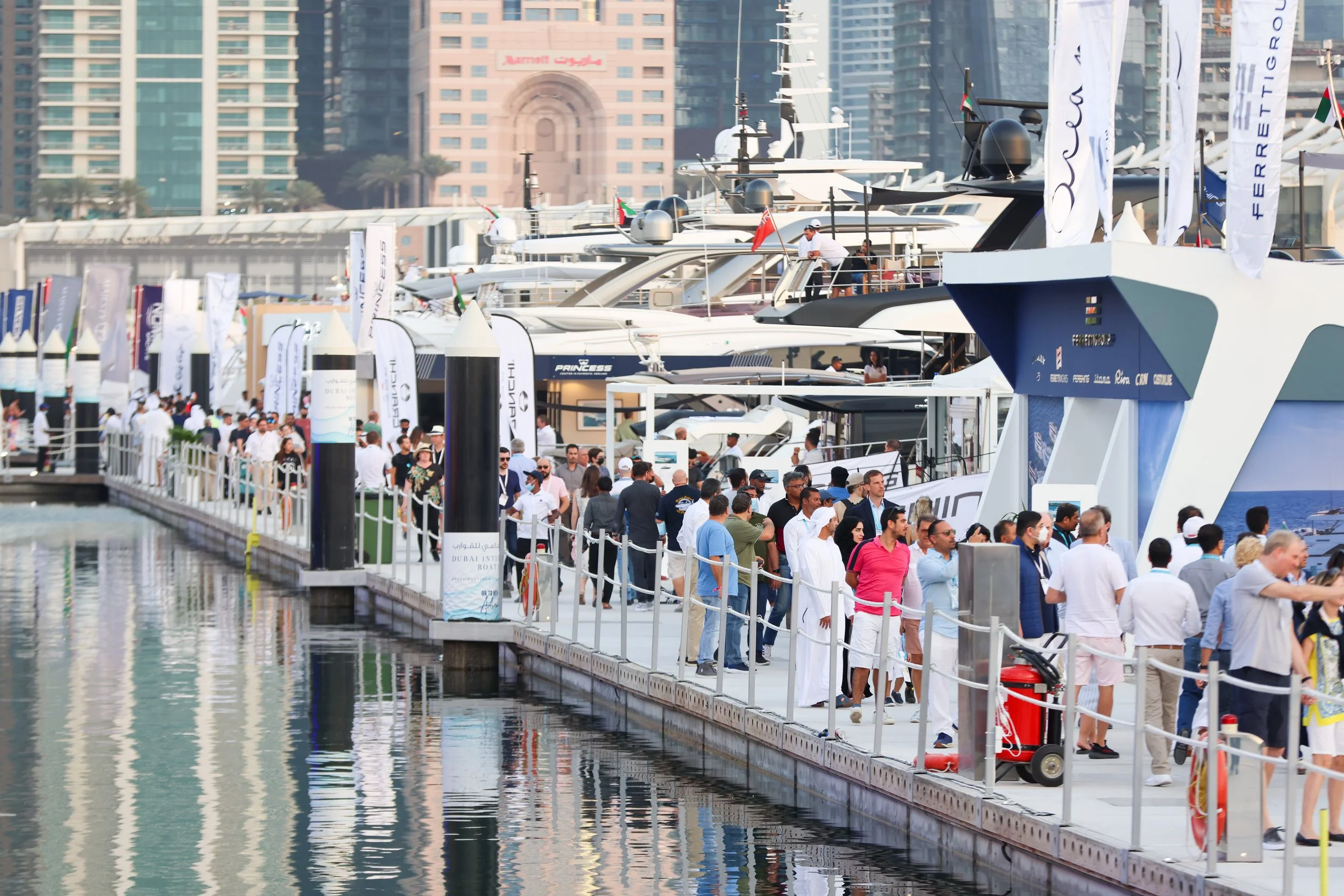 Crowd of people walking along a marina with yachts and high-rise buildings in the background.