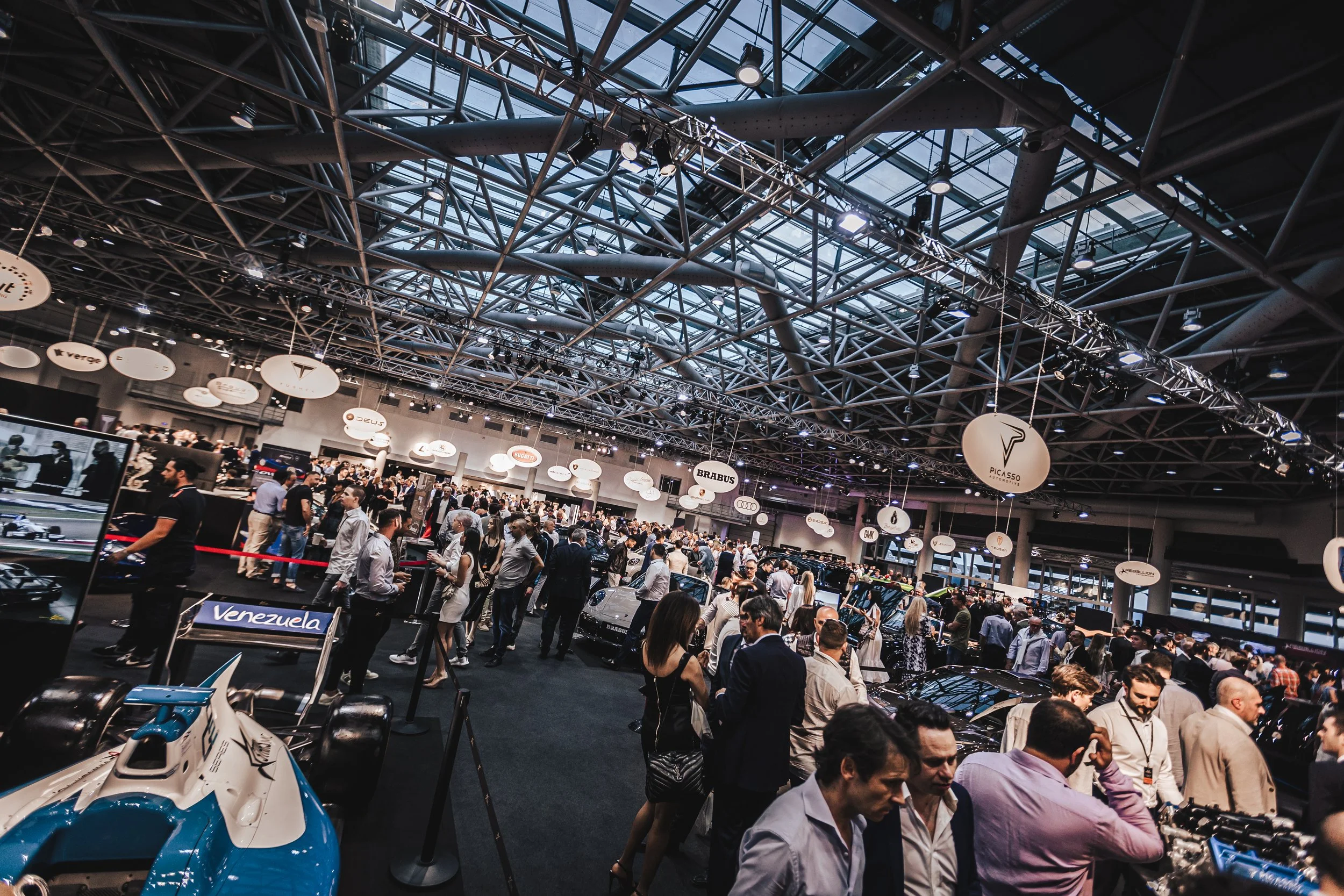 A busy indoor automotive exhibition with numerous cars, people, and automotive brand logos hanging from the ceiling.