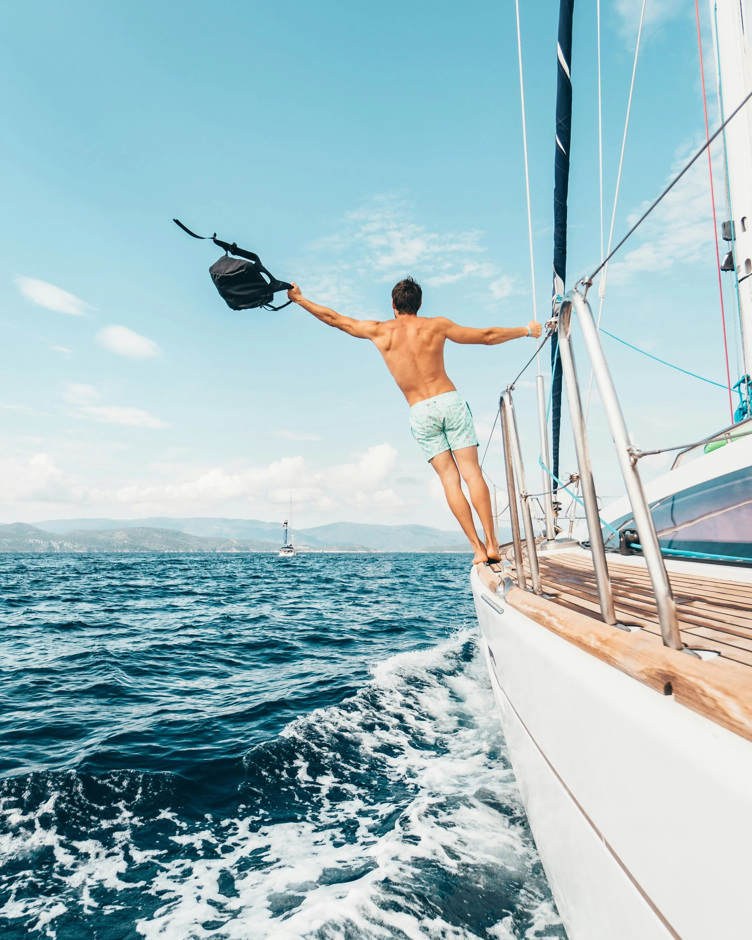 A man is jumping off a sailboat into the water with arms extended, holding a backpack in one hand, on a sunny day with a blue sky and clouds.