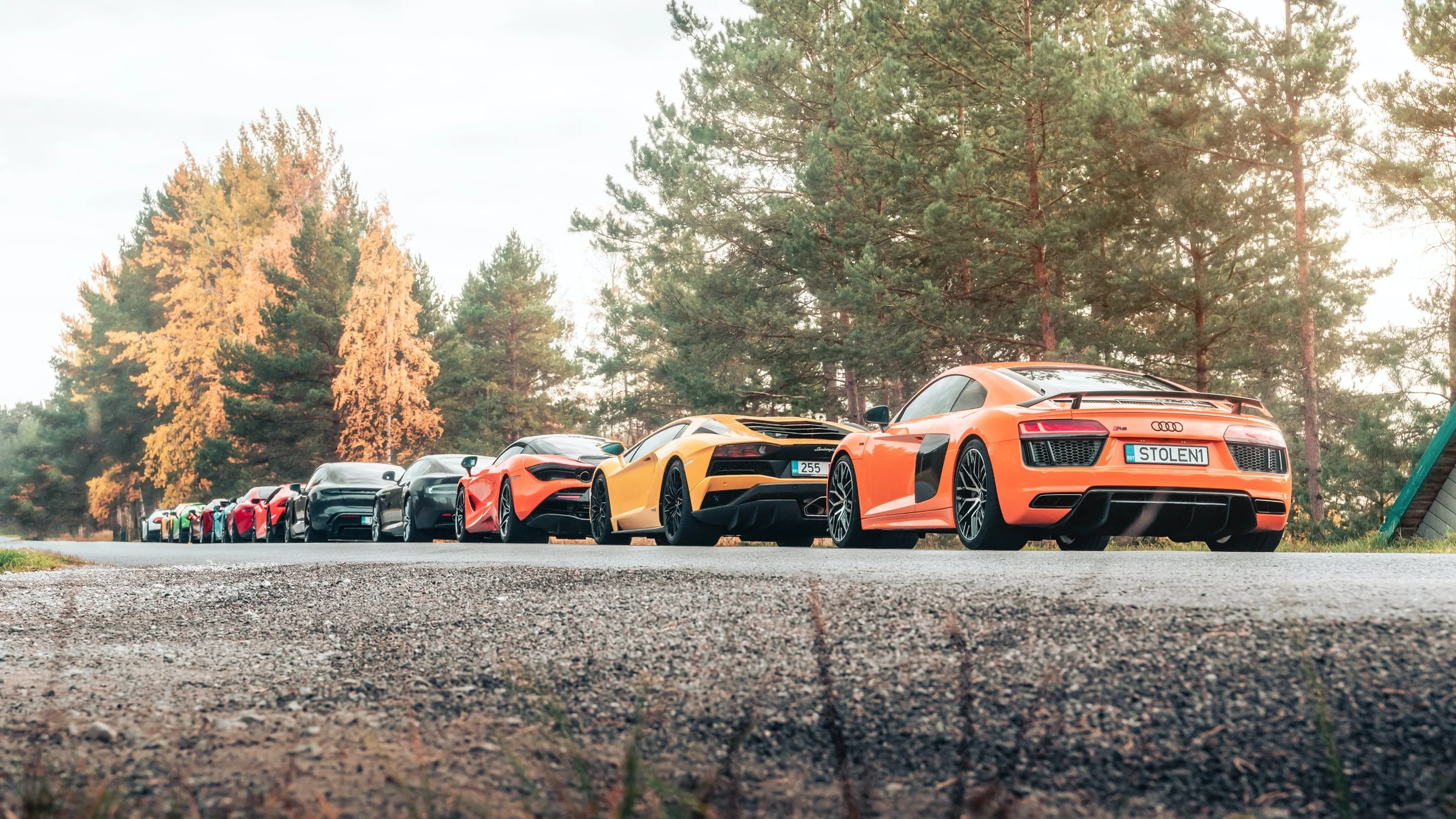 Line of luxury sports cars parked on a rural road with trees in the background during autumn.
