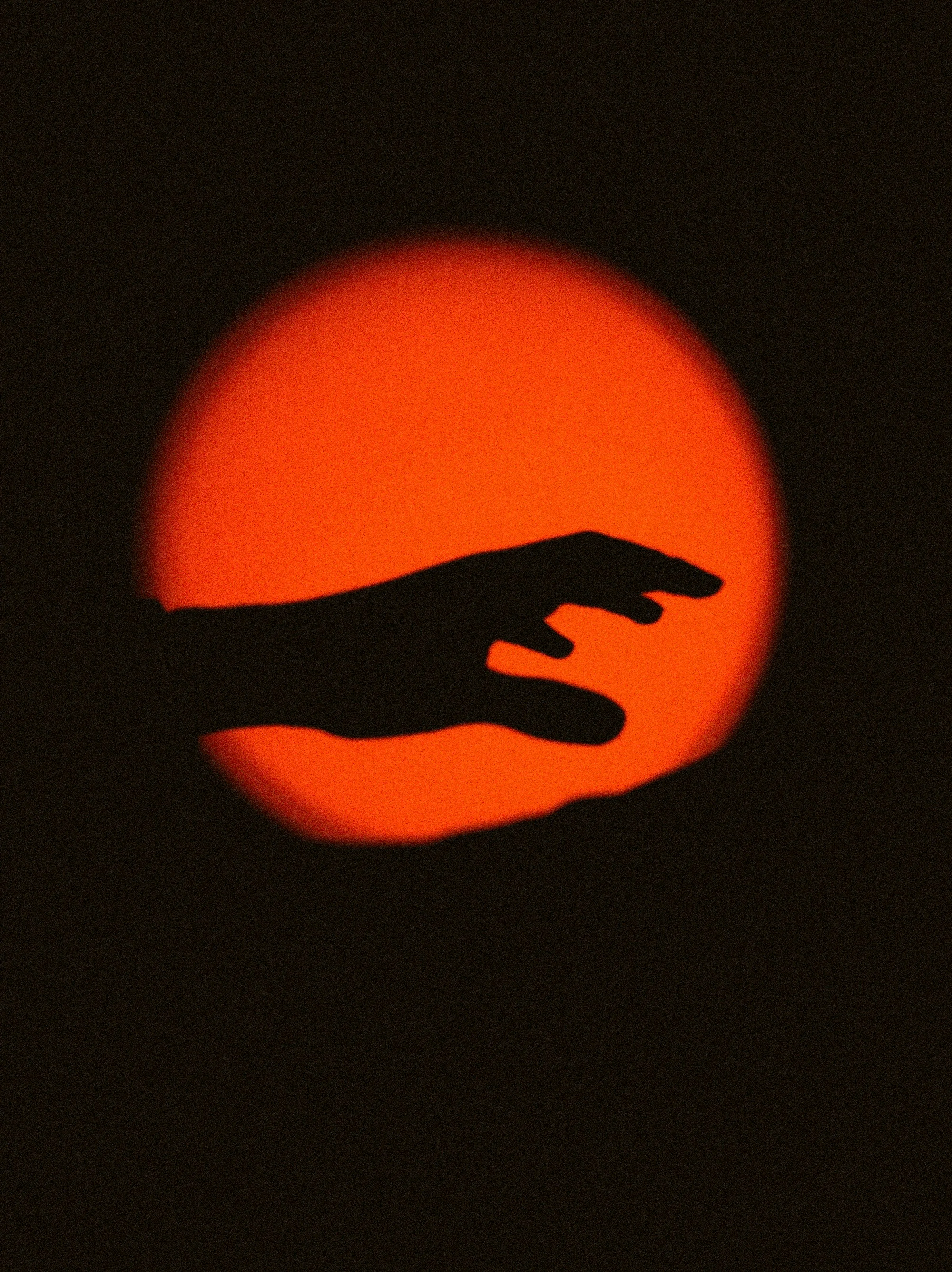 Silhouette of a hand reaching towards an orange sun set at dusk.
