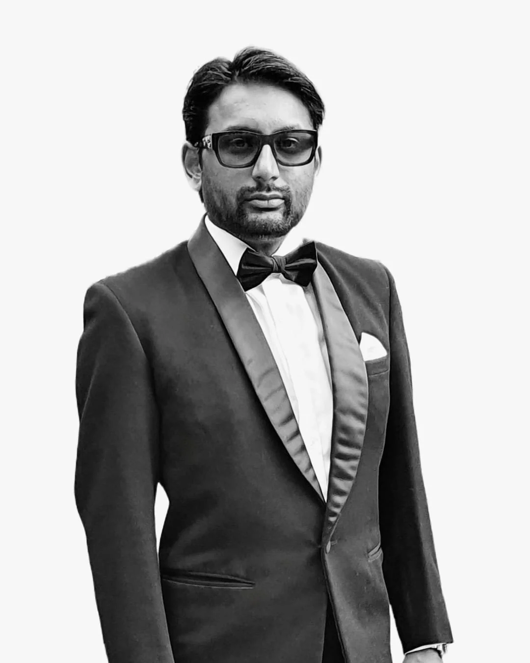 Man dressed in formal tuxedo with bow tie, glasses, and pocket square, standing against a plain background.