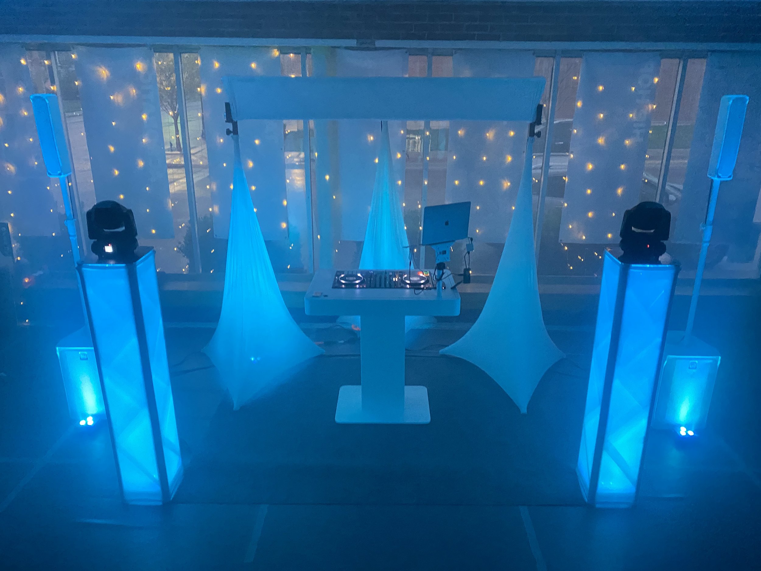 A DJ booth setup with blue lighting, featuring two light towers, a DJ controller, and a laptop on a stand, with decorative white fabric shapes in the background illuminated by string lights.