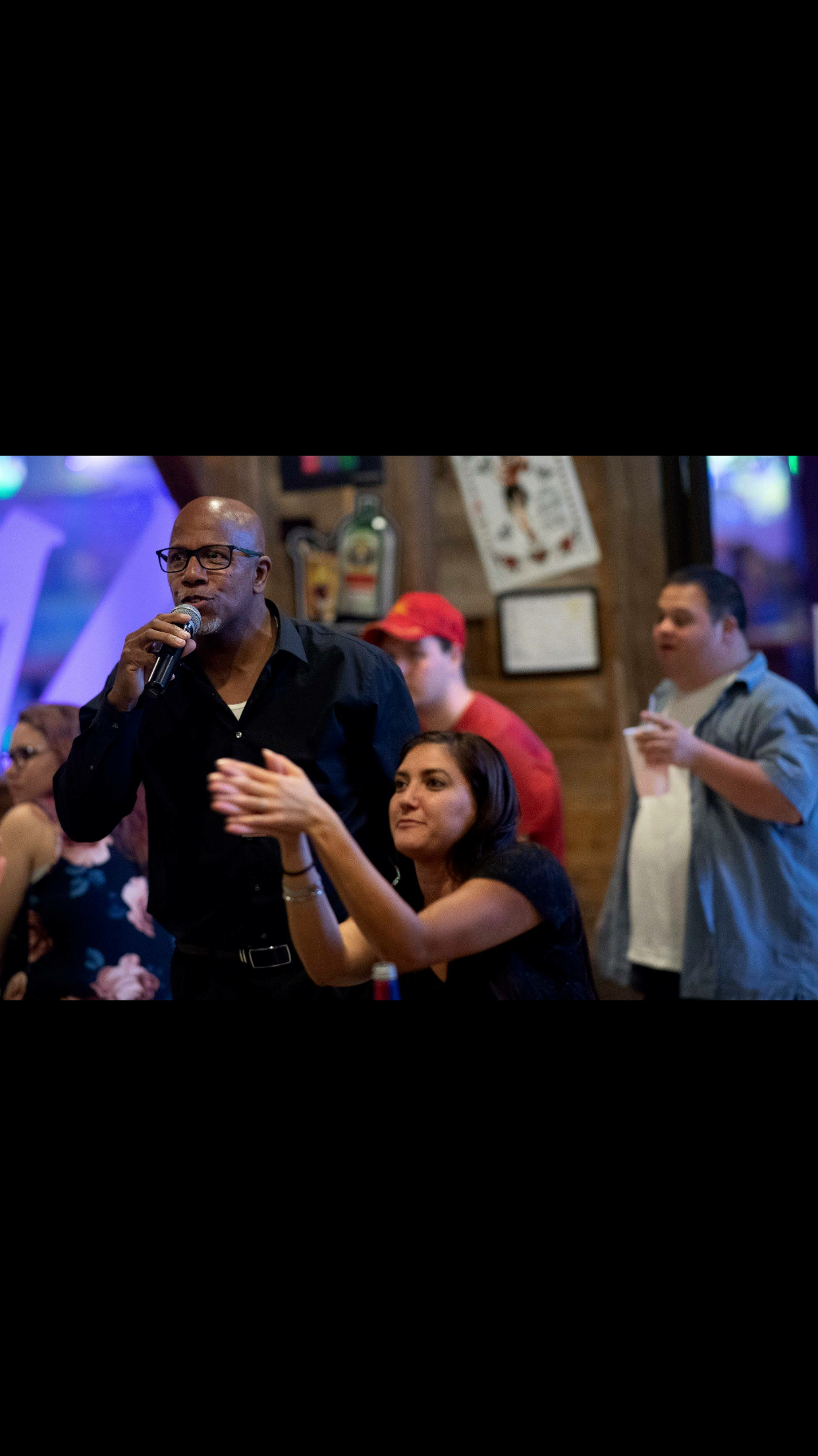 A man with glasses holding a microphone speaks at a crowded bar or restaurant, with a woman clapping in front of him. Other people are in the background, including a man in a red cap and a man in a blue shirt drinking from a cup. The setting has wooden walls and framed pictures.