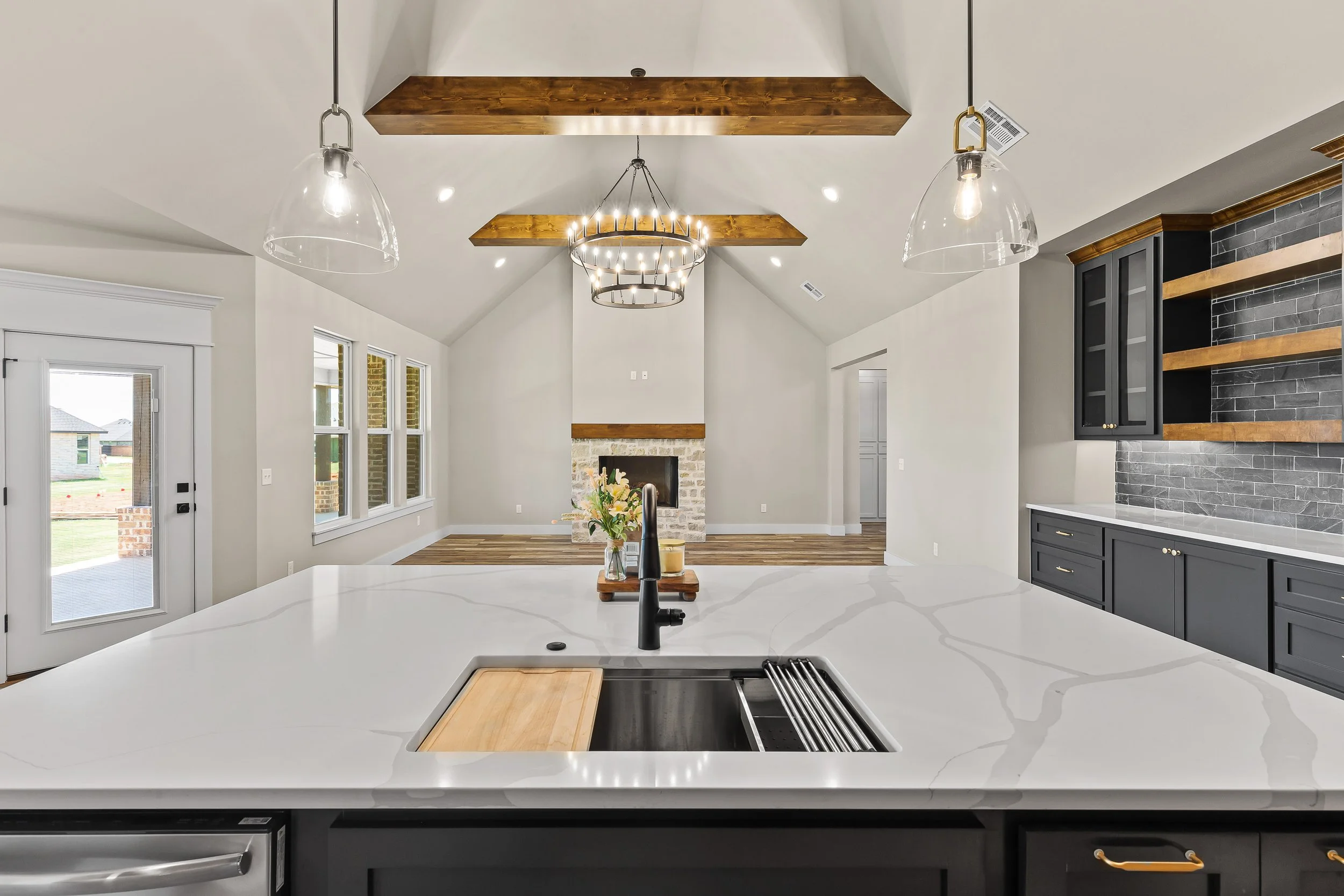 Modern kitchen with white marble countertops, black and gray cabinetry, a gray subway tile backsplash, hanging pendant lights, and a view of a living room with a fireplace and wooden ceiling beams. Real estate photography in Mustang, OK.