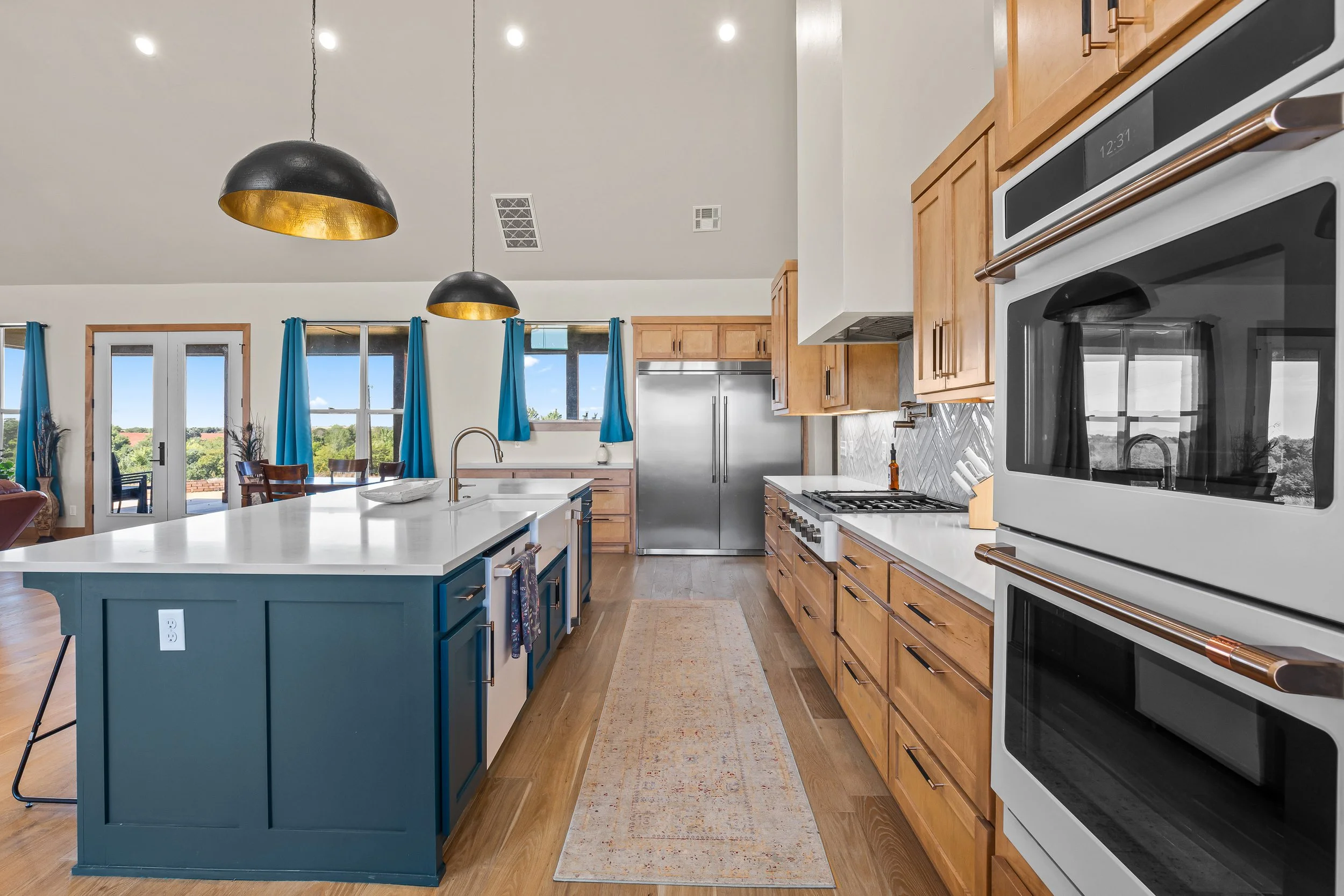 Modern kitchen with wooden cabinets, stainless steel appliances, and a kitchen island painted blue, decorated with pendant lights and a view of a backyard through large windows and a sliding door. Real estate photography in Geary, OK.