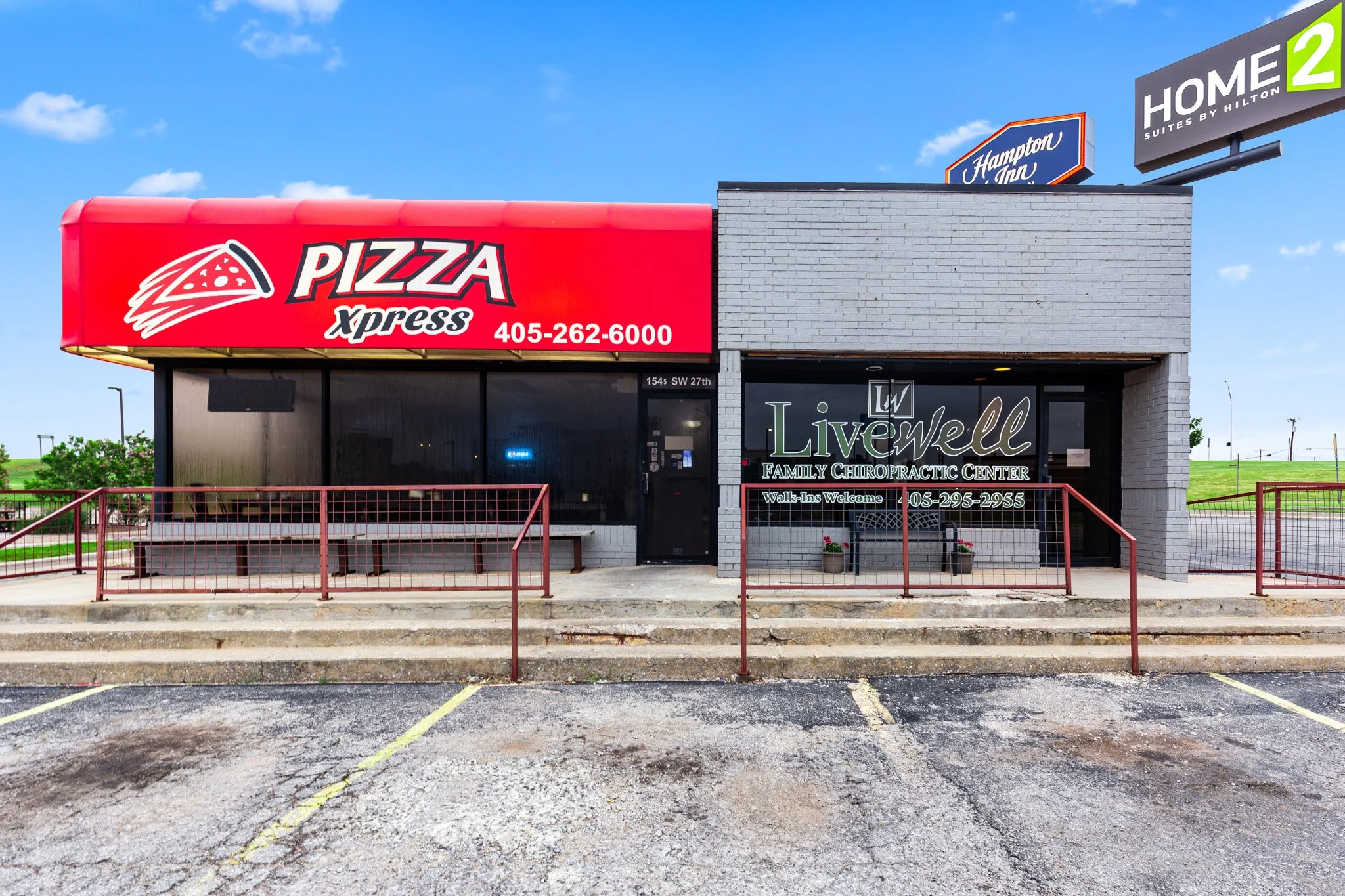 Exterior view of a small commercial building with a bright red sign that reads Pizza Xpress and displays a phone number, and a gray brick facade. Adjacent to it, there is a sign for Livewell Family Chiropractic Center, cautioning walk-ins are welcome